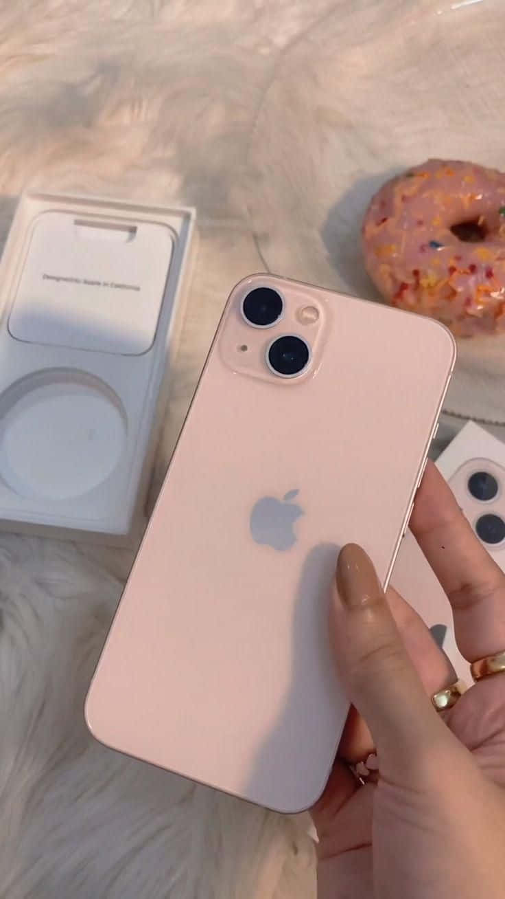 「體驗最新科技，感受經典 iPhone 12」 壁紙 — 一隻手拿著玫瑰金色 iPhone 13 mini，配以冬甩和 MagSafe 充電器，放在毛絨白色表面上。