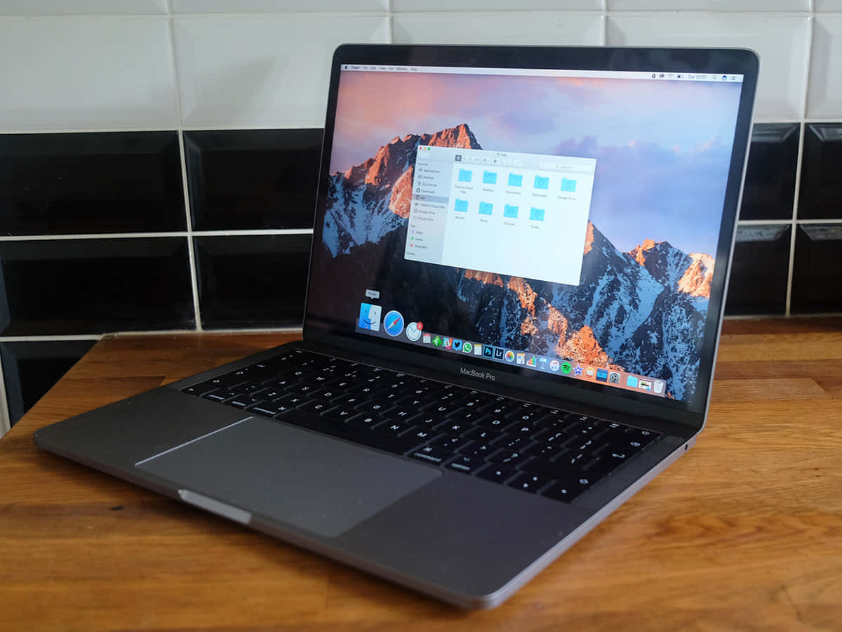 體驗流暢效能，選擇時尚優雅的 MacBook 壁紙 — 一部銀色 MacBook Pro 打開放在木桌上，背景係黑色瓷磚牆，屏幕上顯示山脈壁紙。