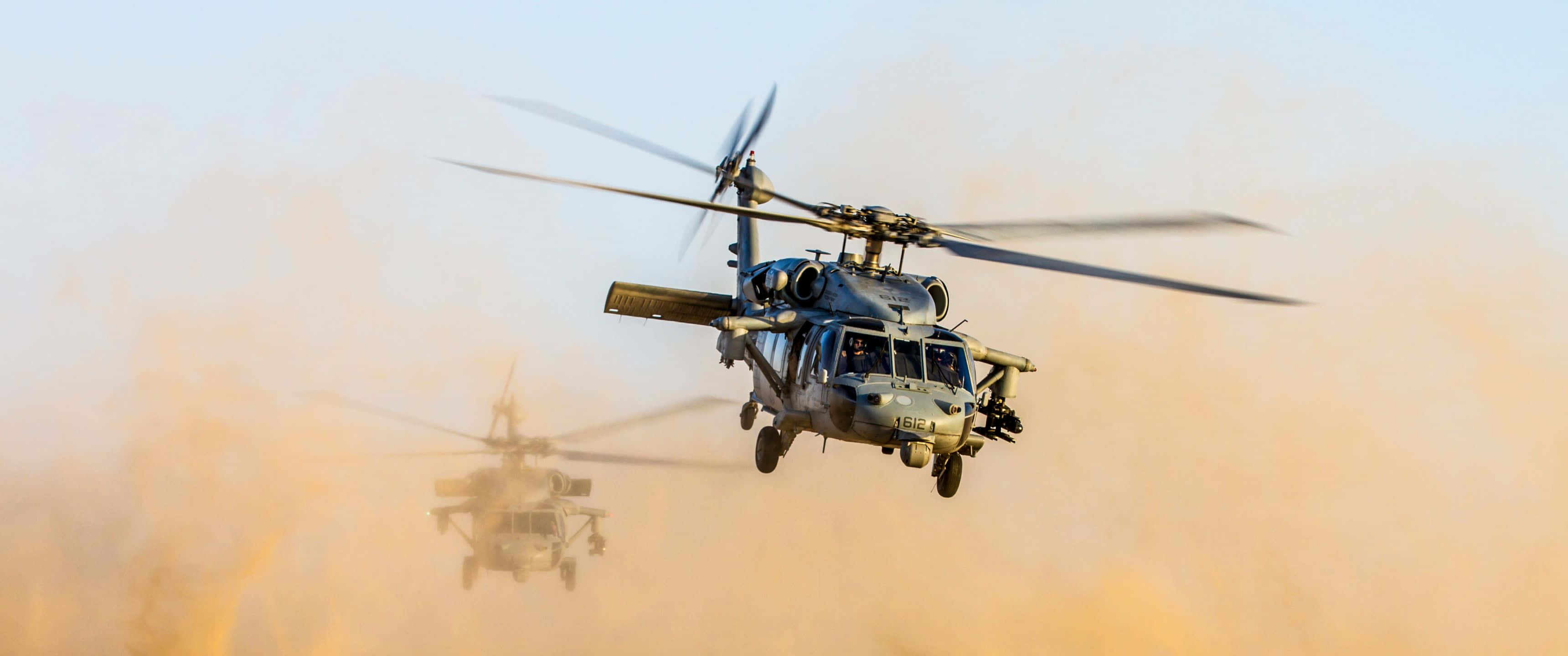 體驗直升機飛行的驚險快感 壁紙 — 兩架軍用直升機低飛穿過塵霧，旋翼槳葉因運動而模糊。