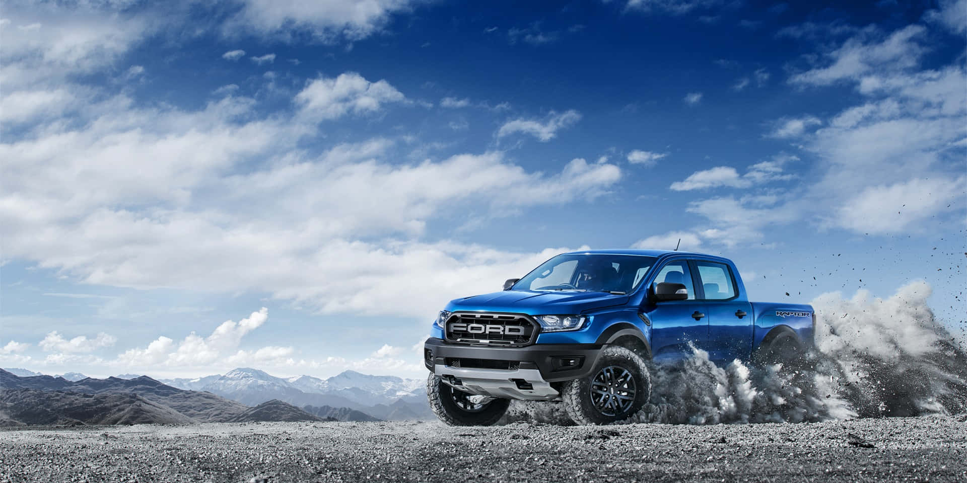 藍色福特 Ranger Raptor 卡車在礫石上行駛，背景有山和雲。