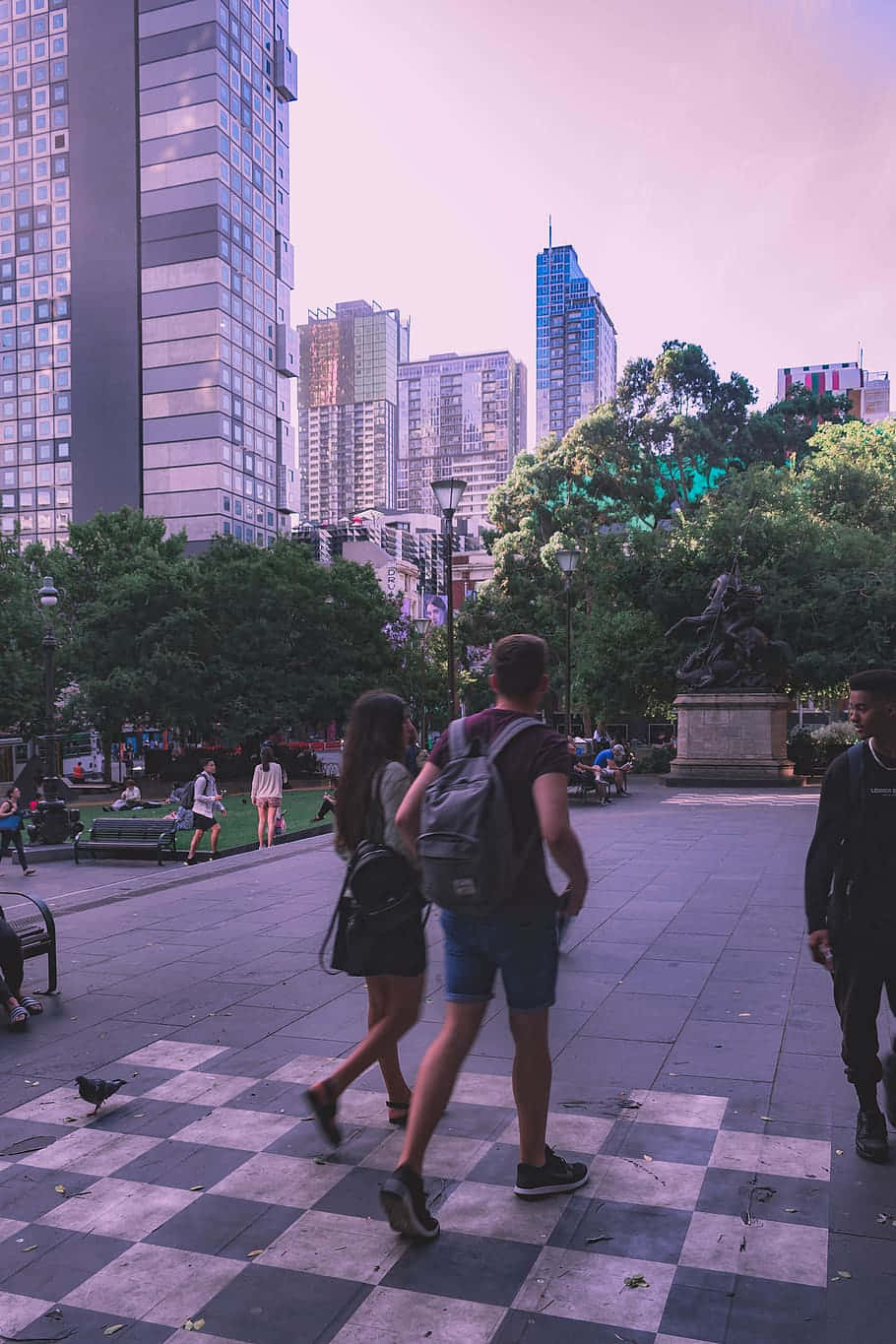 日落時分，人們在棋盤格廣場上行走，背景是城市建築和樹木。