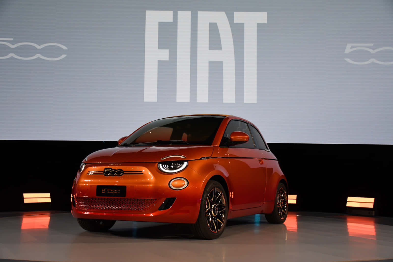 體驗菲亞特的卓越品質 壁紙 — 一輛充滿活力的橙色Fiat 500e電動車在時尚的汽車發布活動中，在舞台燈光下閃閃發光。