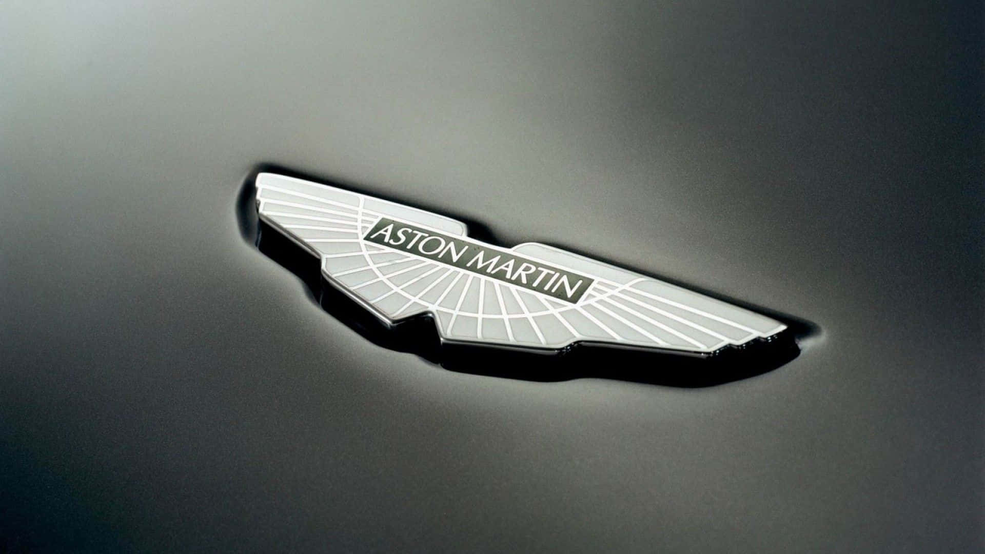 體驗阿斯頓·馬丁的動力、性能與尊貴 壁紙 — 特寫：銀色Aston Martin翼形標誌徽章，置於時尚深色引擎蓋上，光線柔和。