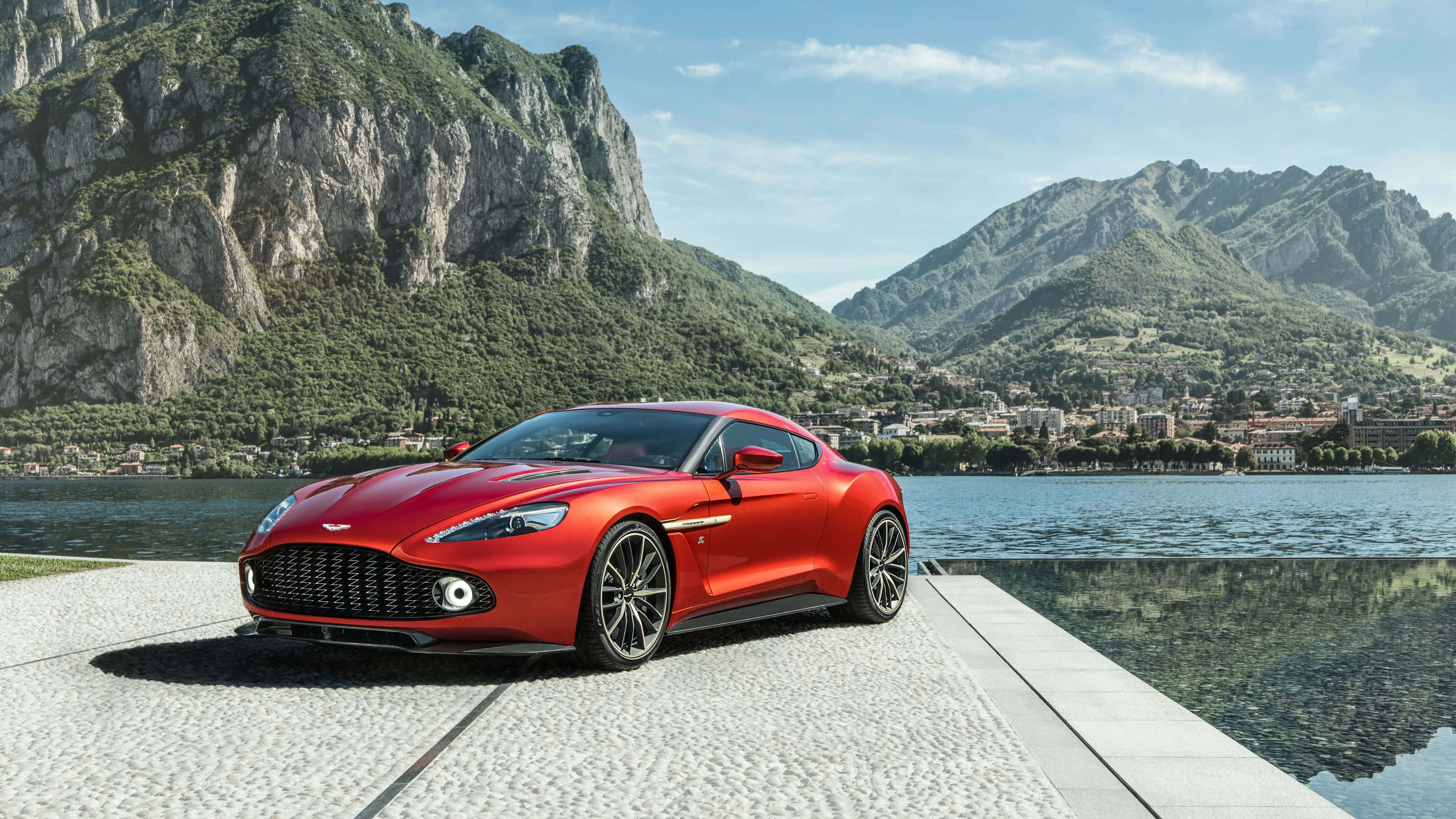 體驗阿斯頓·馬丁的奢華 壁紙 — 一輛鮮紅色的Aston Martin跑車在湖邊碼頭上閃閃發光，背景是崎嶇的山脈和一個小鎮，在清澈的藍天下。
