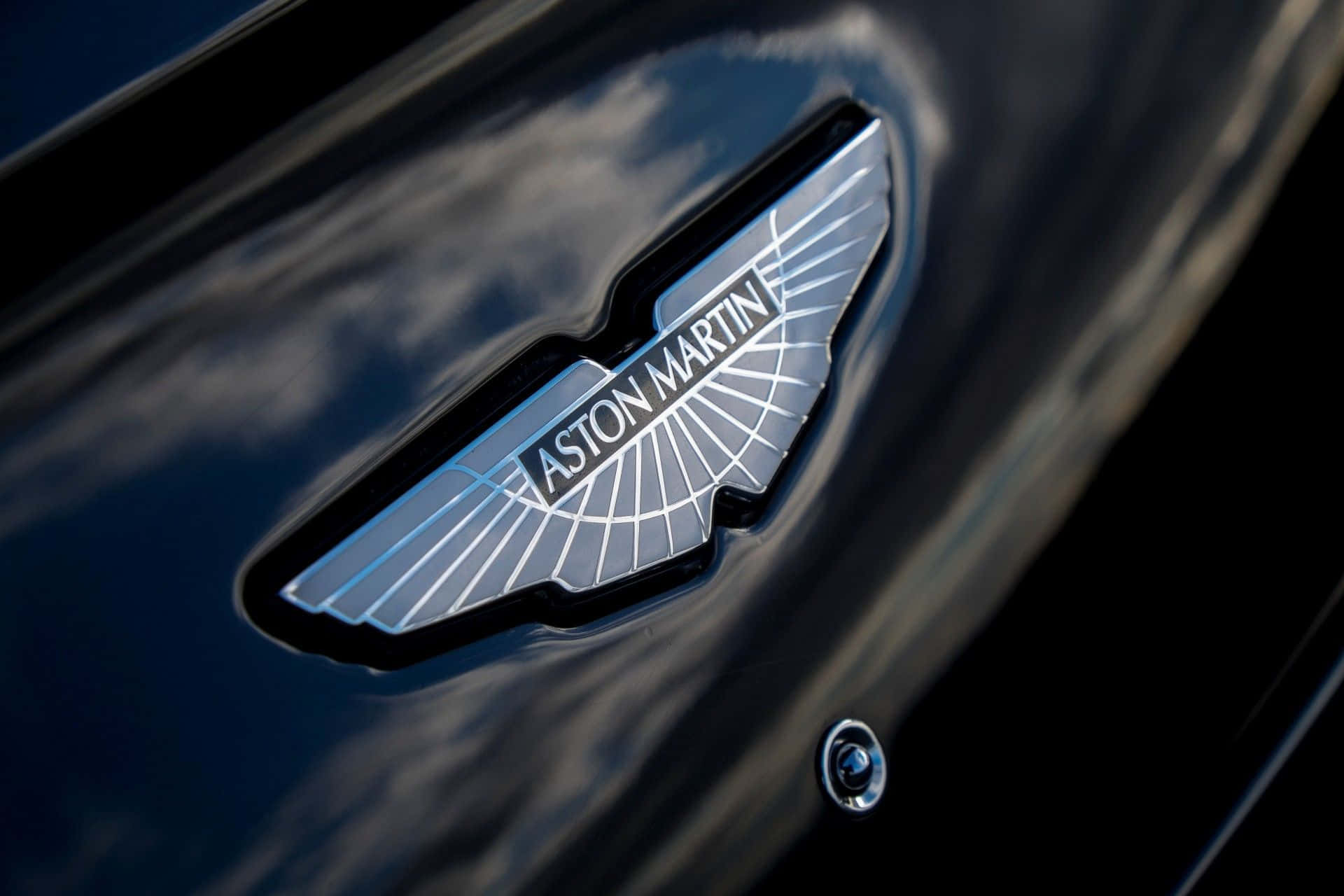 體驗阿斯顿·马丁的奢華駕馭樂趣 壁紙 — 特寫：閃亮銀色Aston Martin翼形標誌，位於反射陰天雲層的光澤黑色引擎蓋上。