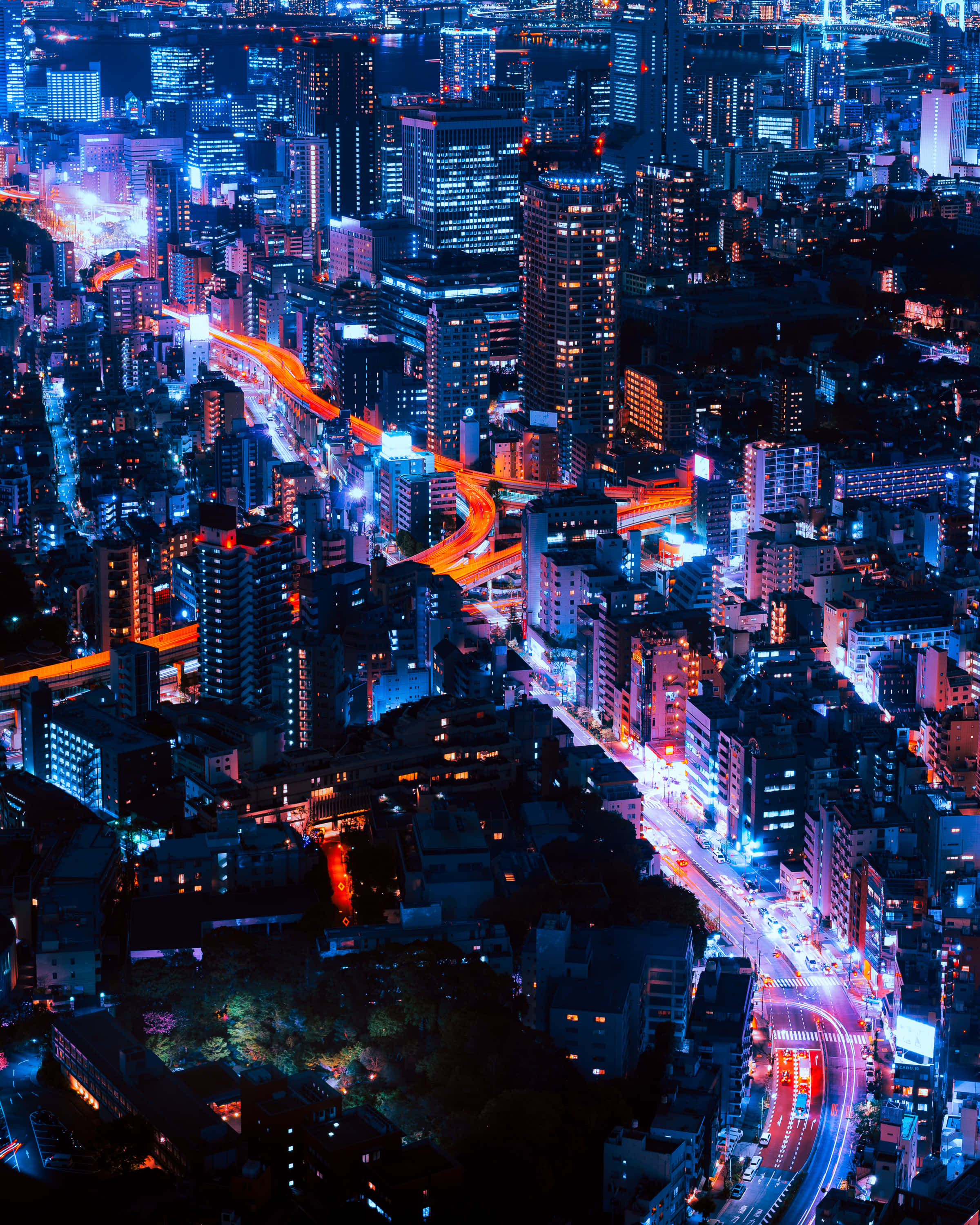 體驗霓虹城市的熱鬧能量 壁紙 — 夜晚霓虹燈照亮城市景觀的鳥瞰圖，交通流動穿過密集摩天大樓時發出藍色和橙色的光軌。