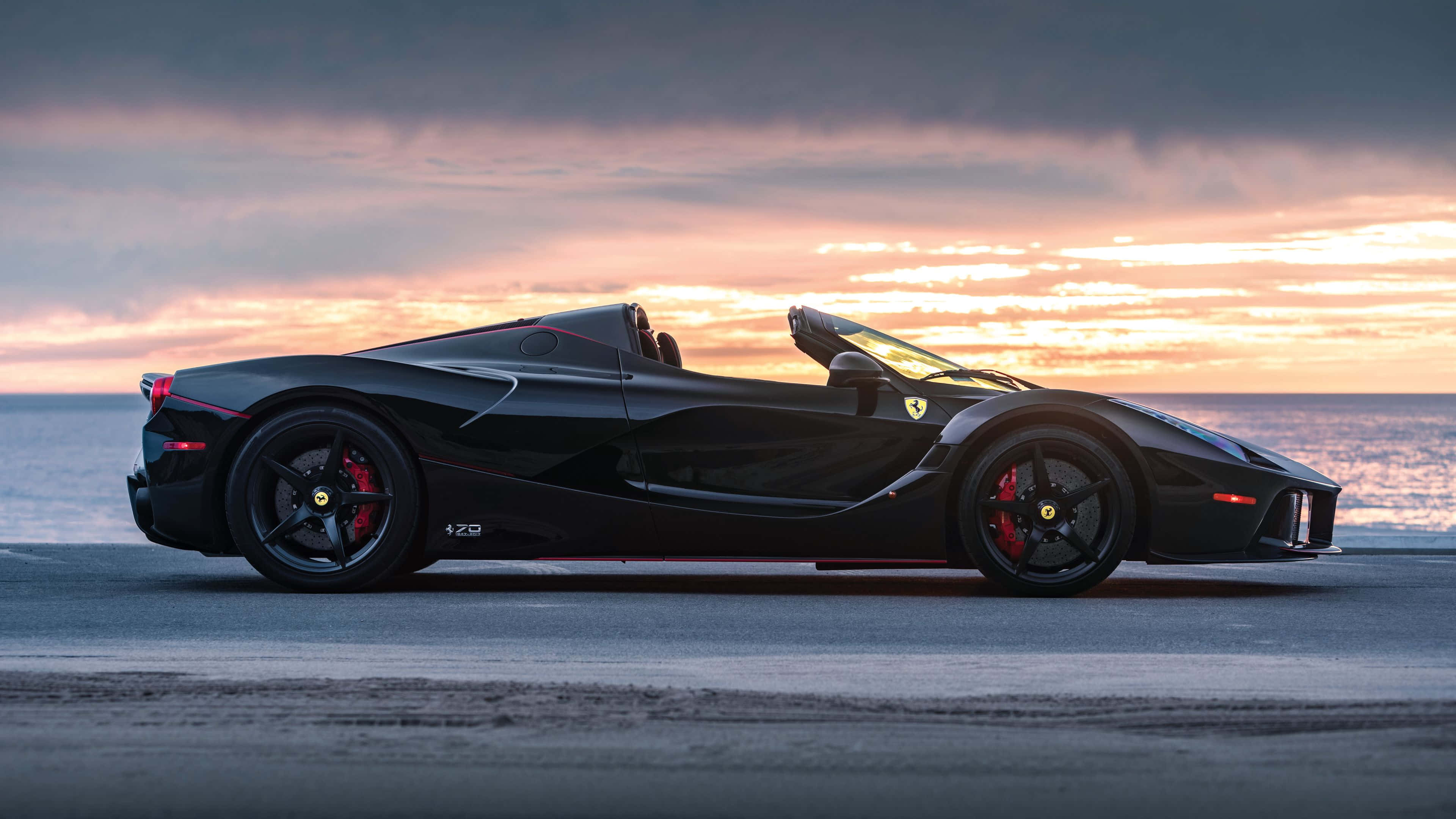 體驗駕駛經典法拉利的狂野快感 壁紙 — 一輛時尚的黑色法拉利 LaFerrari Aperta 停泊在日落時分的海岸公路上，海洋上空有鮮豔的橙色和粉紅色雲彩。