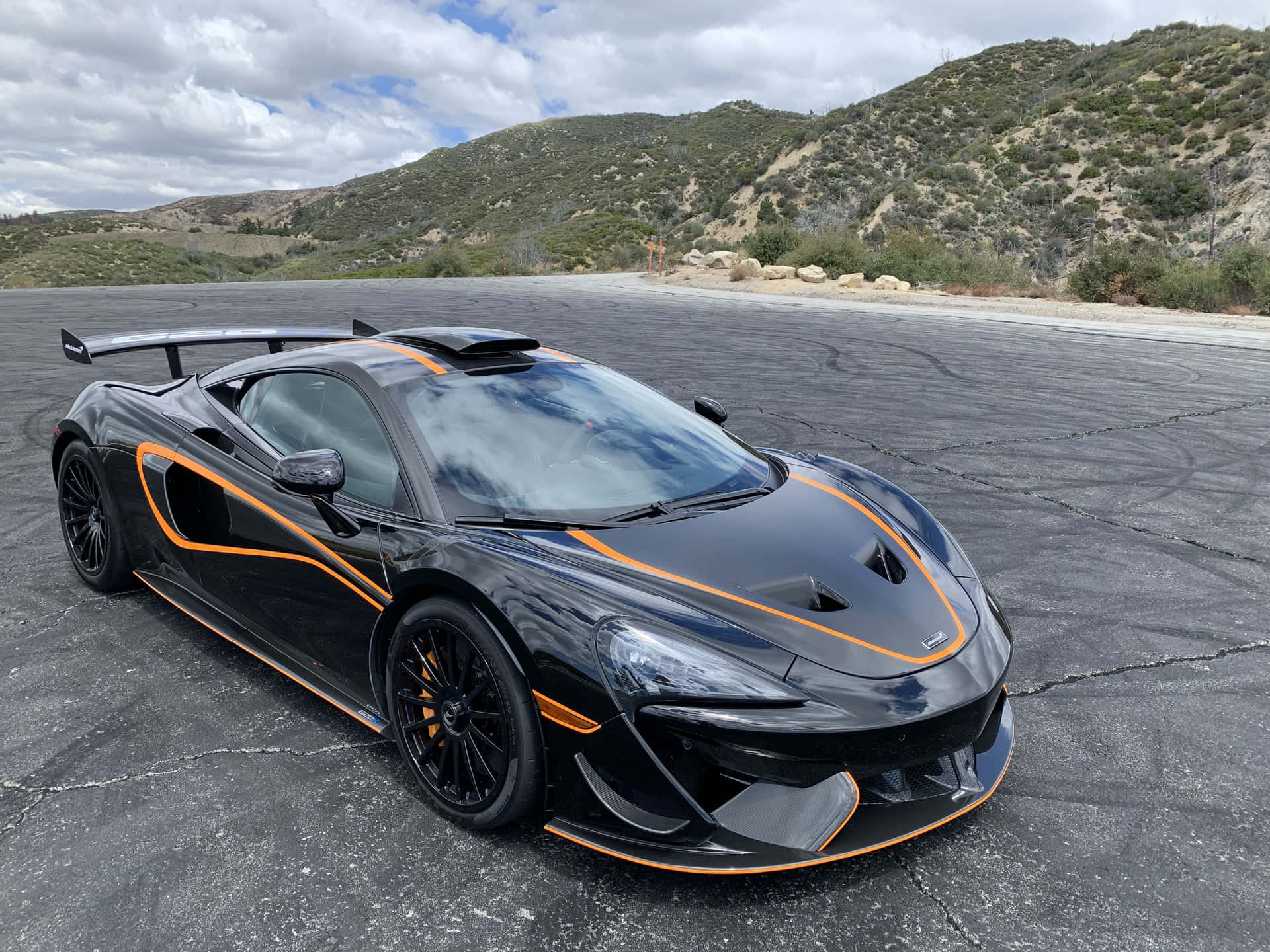 黑色McLaren跑車帶橙色飾邊停在瀝青地上，靠近山丘。