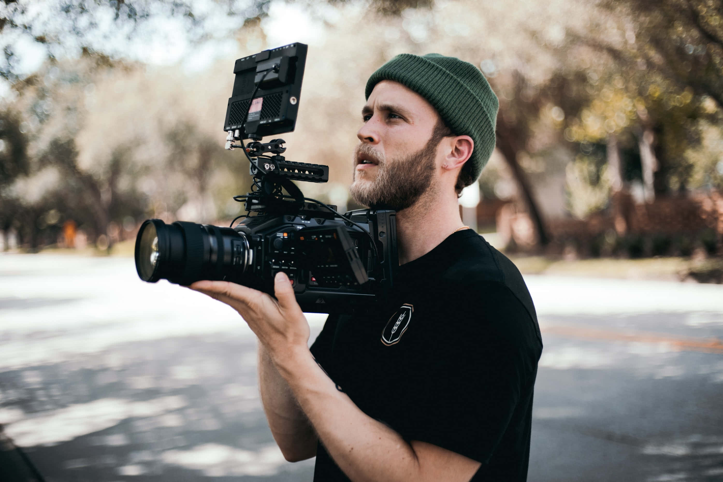 高品質 DSLR 相機 壁紙 — 一位留鬍鬚的男子戴著綠色無簷帽，手持帶有顯示器和鏡頭的專業相機，站在戶外綠樹成蔭的街道上。