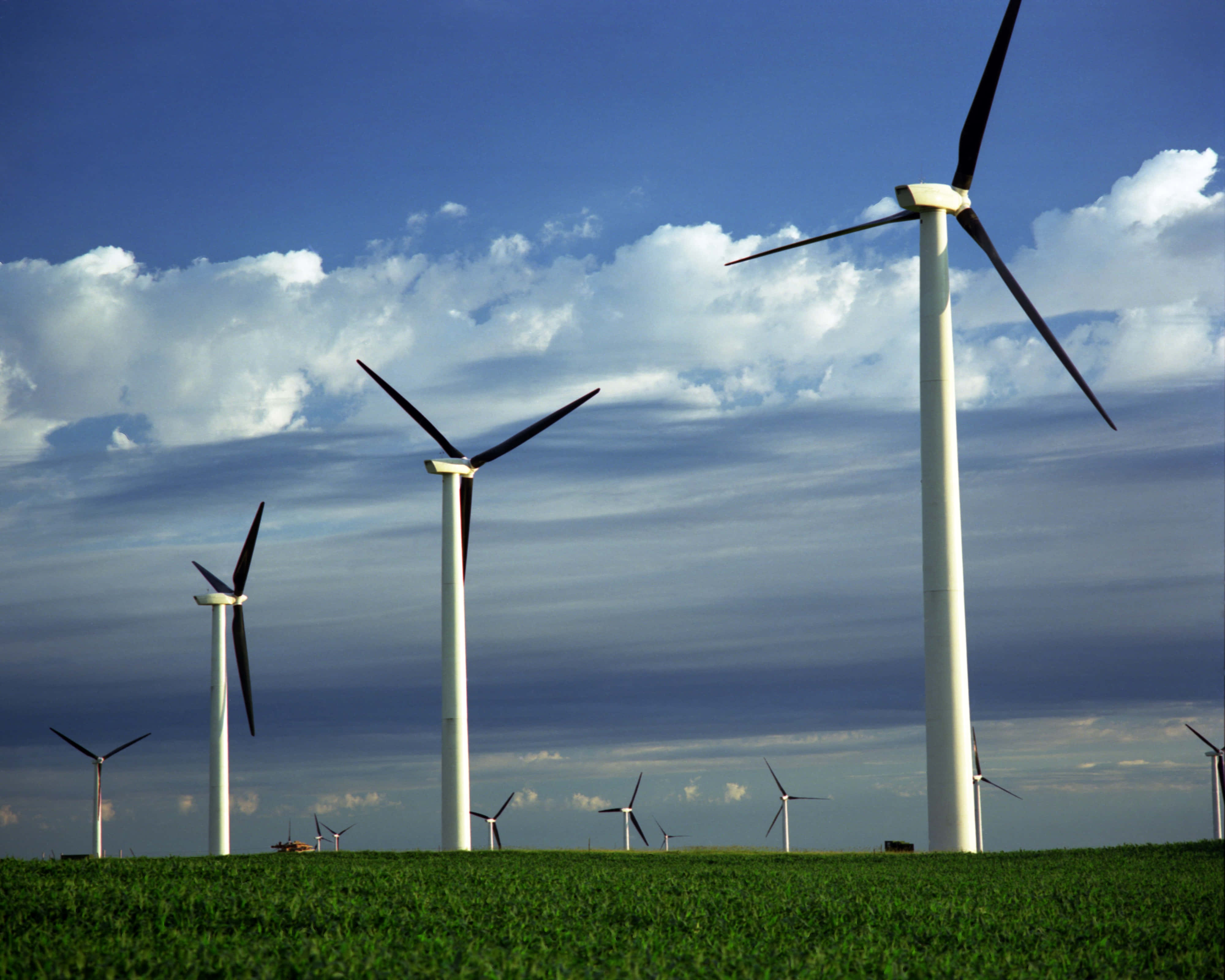 Energy 桌布 壁紙 — 高大的白色風力渦輪機矗立在充滿活力的綠色田野中，上方是戲劇性的藍天，點綴著蓬鬆的白雲。