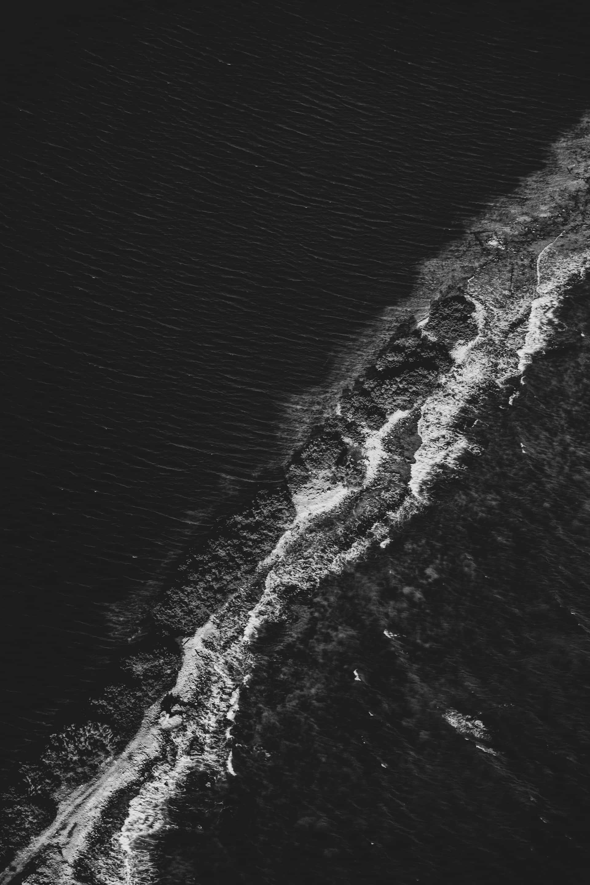 Black And White Ocean 桌布 壁紙 — 高對比度黑白航拍照片，展示海浪拍打黑暗岩石海岸線的景象。