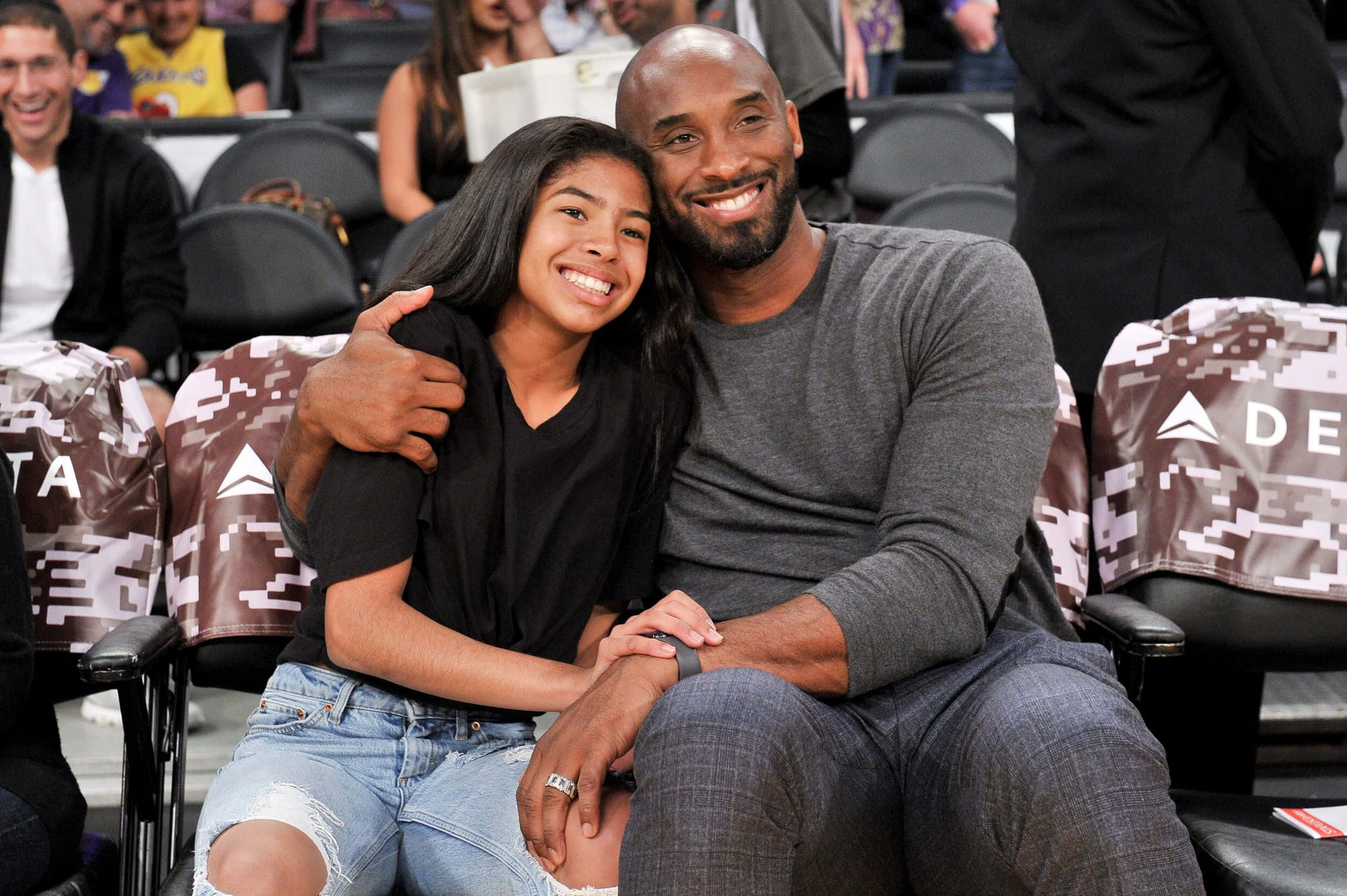 Kobe And Gigi 桌布 壁紙 — 高比拜仁與女兒吉安娜在籃球比賽中坐在一起，背景是體育館座位和球迷，溫暖地微笑。