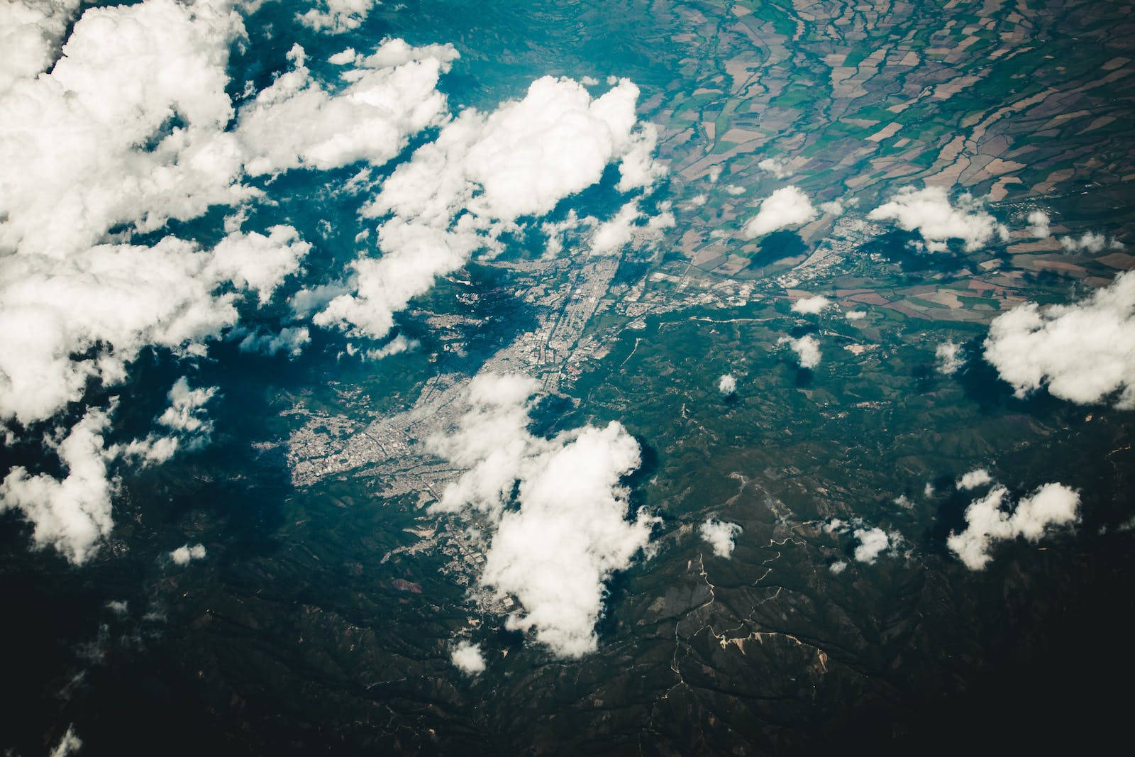 高海拔地形 壁紙 — 俯瞰一座廣闊城市，坐落於翠綠山丘與農田之間，部分被蓬鬆白雲遮蔽。