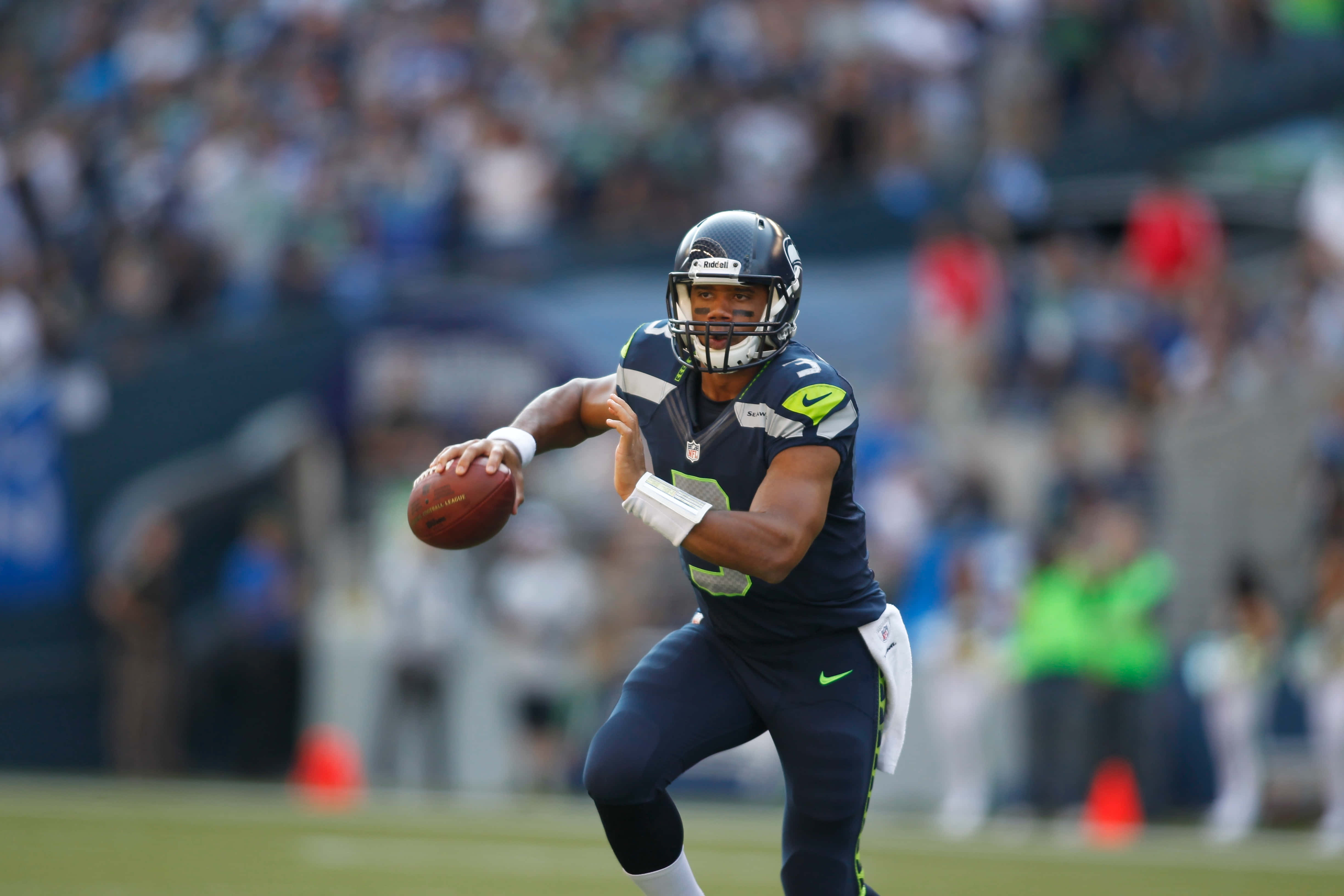 高清NFL壁紙，感受美國最愛運動的熱情 壁紙 — 一位身穿海軍藍和綠色球衣的西雅圖海鷹隊四分衛在日間比賽中擲出橄欖球，背景是模糊的體育場人群。