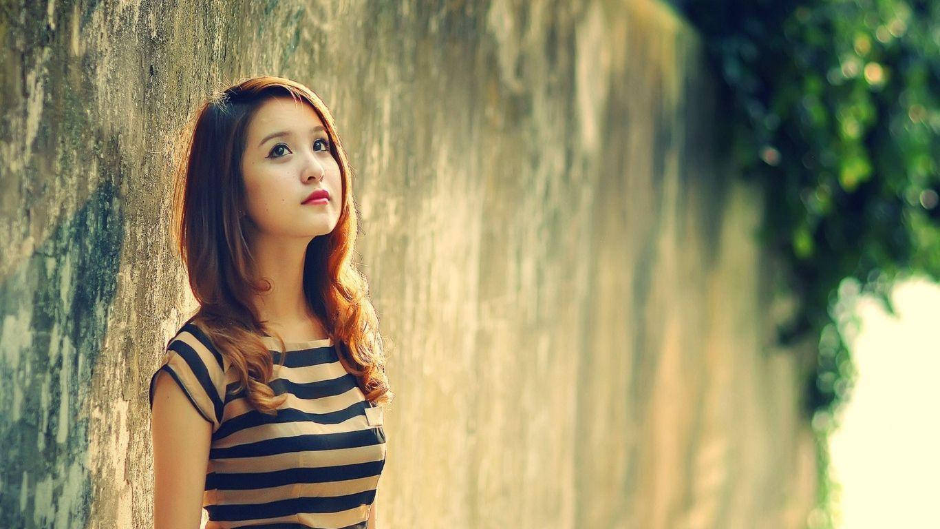 高清女孩 抬頭姿勢 壁紙 — 一位身穿條紋上衣的年輕女子靠著風化的石牆，背景中的綠植柔和地模糊。