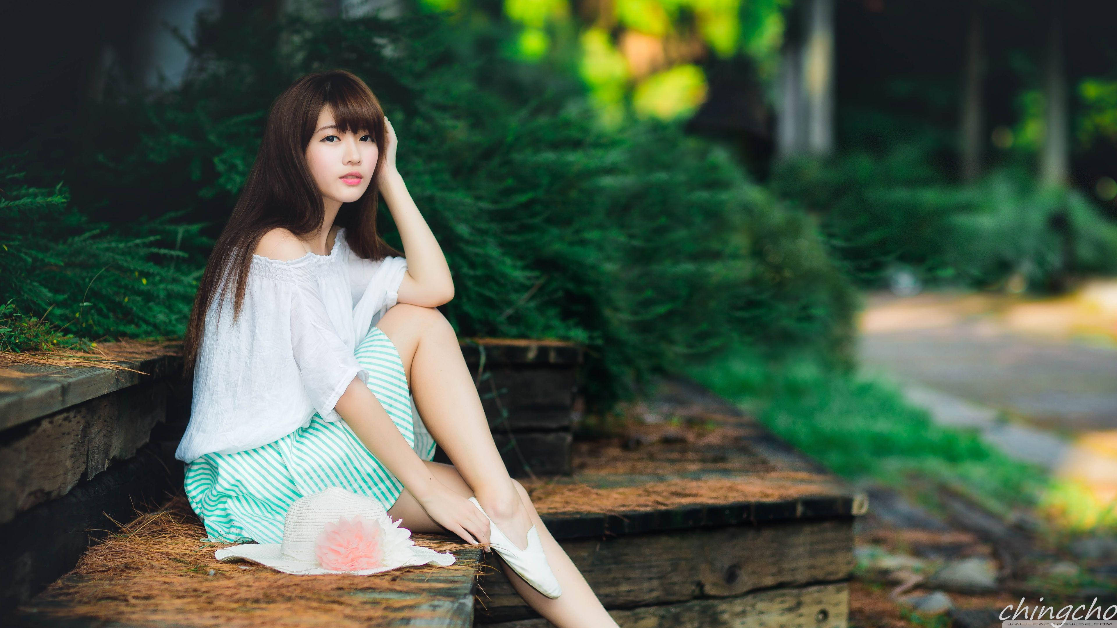 高清女孩亞洲模特 壁紙 — 一位身穿白色上衣和條紋裙的年輕女士優雅地坐在木階梯上，周圍綠意盎然。