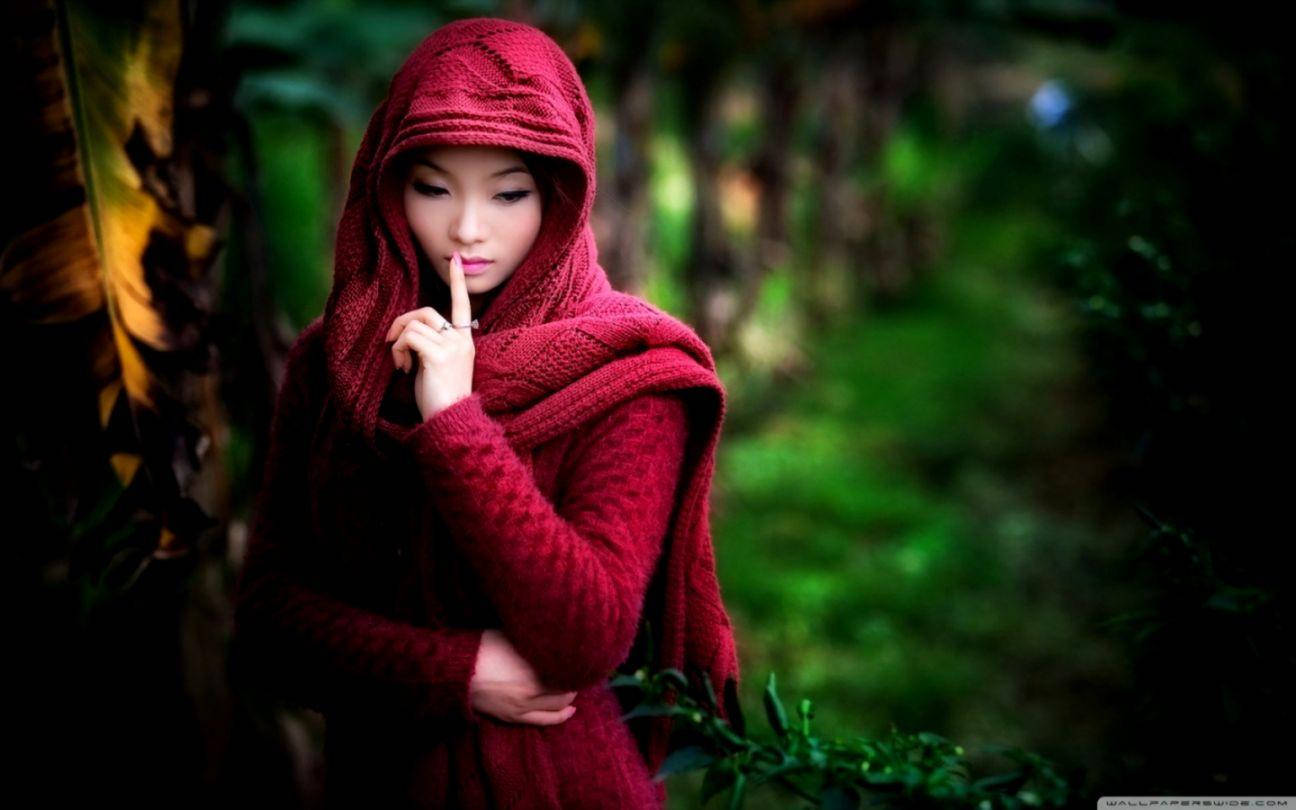 高清女孩戴頭巾服裝 壁紙 — 一位身穿深紅色針織連帽毛衣的女性站在茂密的綠色森林中，將手指放在嘴唇上做出安靜的手勢。