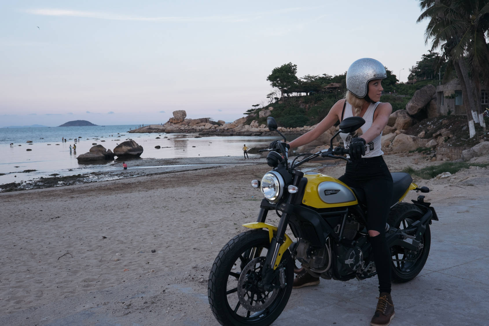 高清女孩與Ducati Scrambler 壁紙 — 一位戴著銀色頭盔的女性坐在一輛停在沙灘上的黃色電單車上，背景是暮色中的棕櫚樹和遠處的岩石。