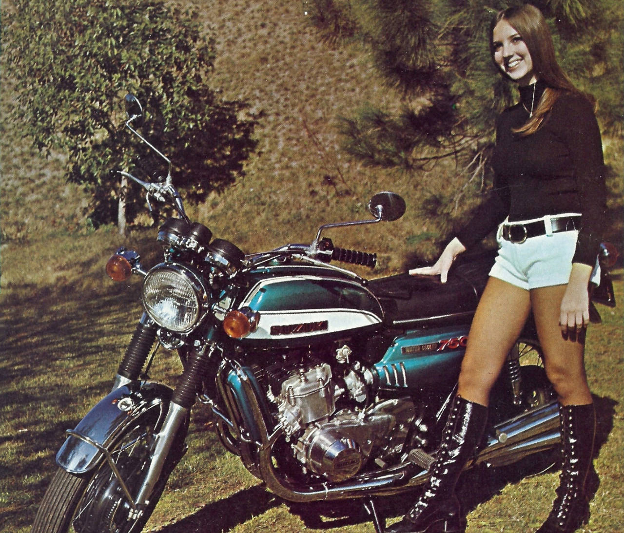 高清女孩與Suzuki摩托車 壁紙 — 一位微笑的女士穿著復古服裝，靠在一輛停在樹下草地上的閃亮青綠色摩托車上。