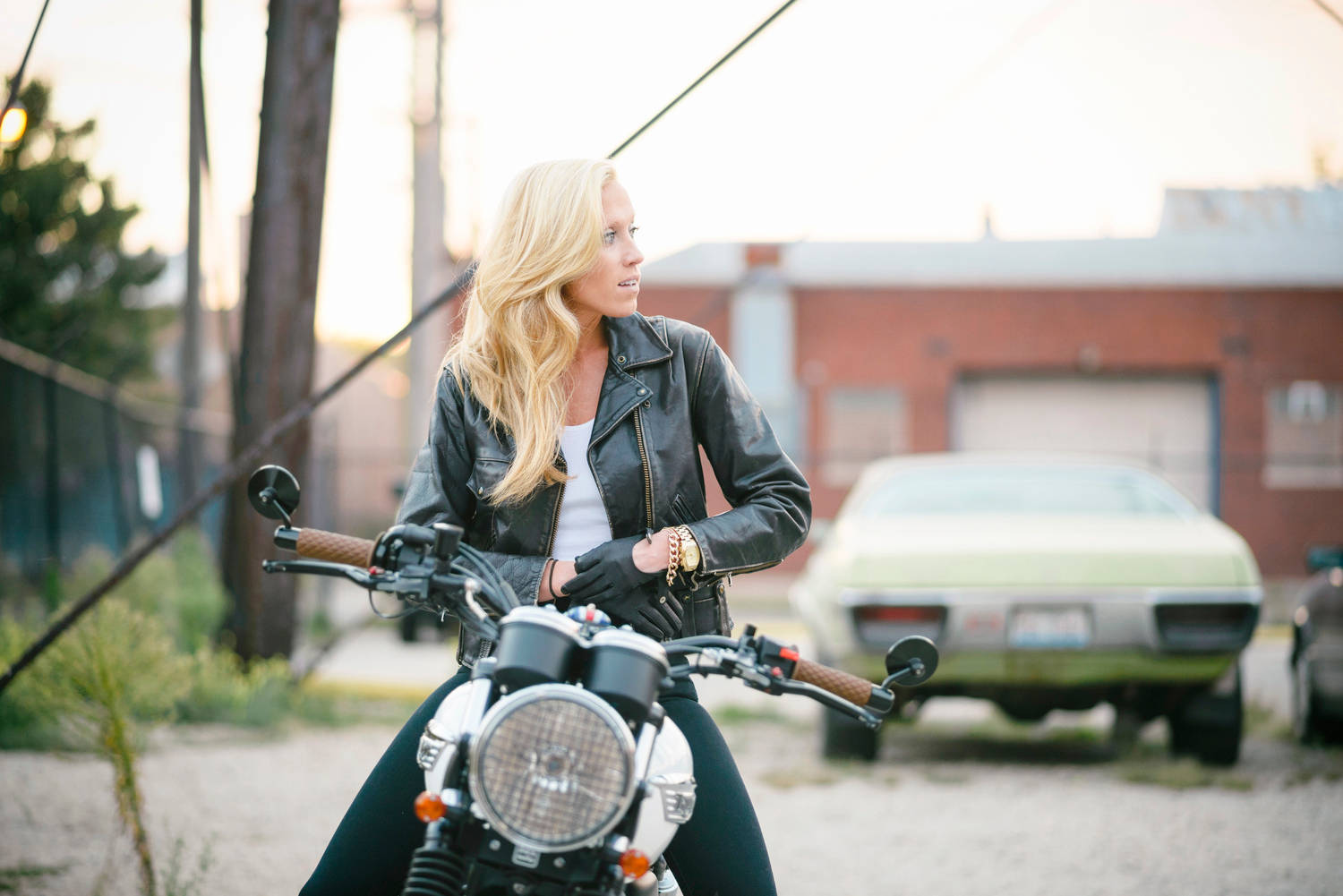 高清女孩與Triumph Bonneville 壁紙 — 一位金髮女子身穿黑色皮夾克，在黃金時段的城市小巷中，優雅地坐在一輛經典摩托車上。