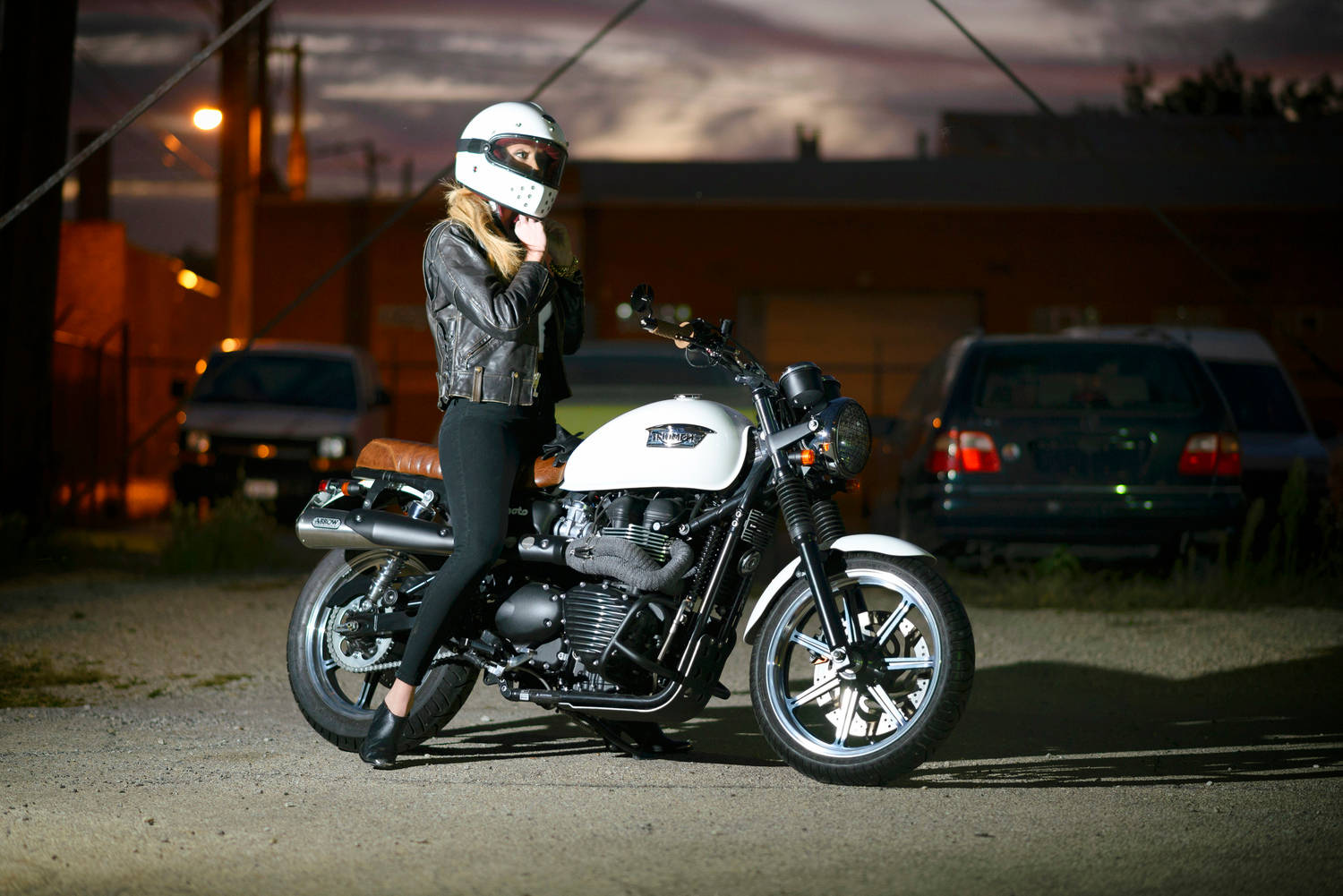 高清女孩與Triumph摩托車 壁紙 — 一位戴著白色頭盔和皮夾克的女性，在暮色天空下，坐在一輛黑白相間的摩托車上，背景中溫暖的路燈閃爍著光芒。