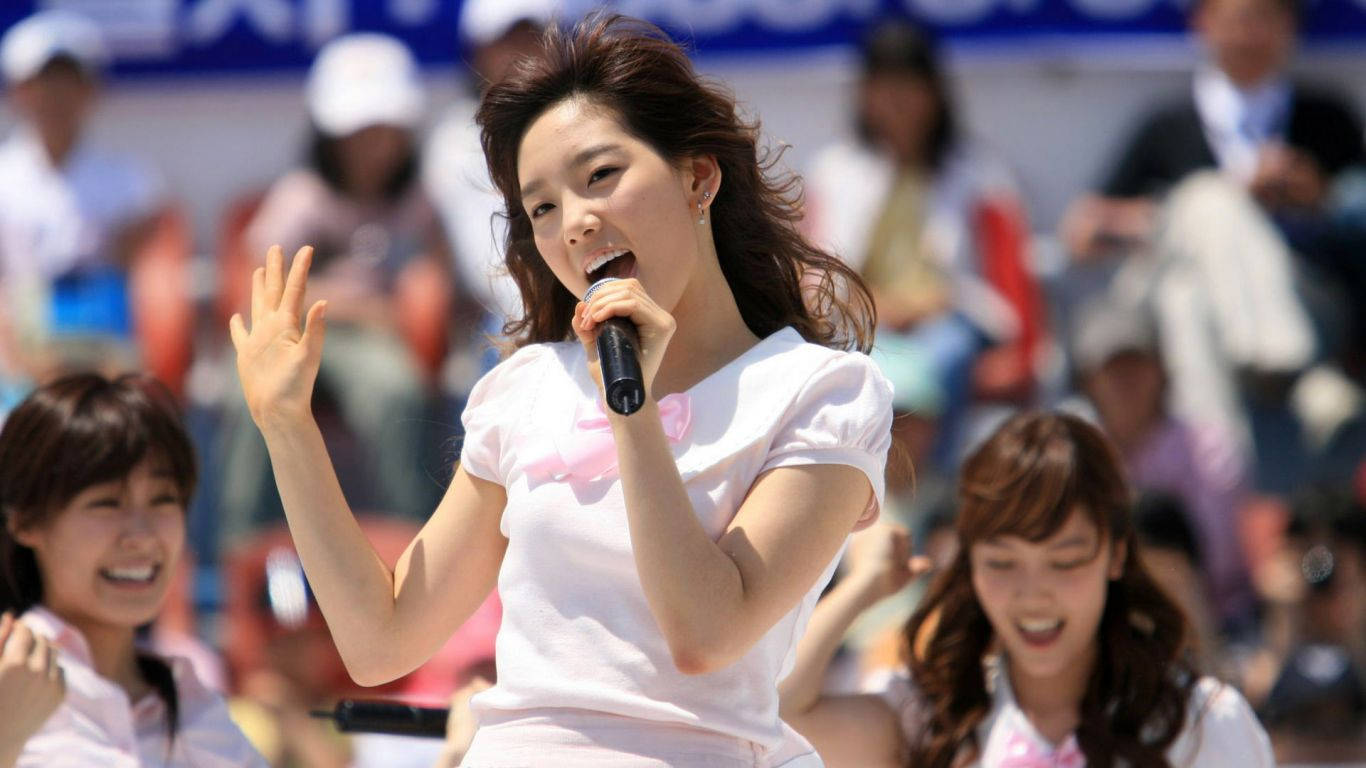 高清少女時代主唱 Tae Yeon 演唱會畫面 壁紙 — 一位微笑的年輕女子穿著白襯衫，在陽光明媚的日子裡對著麥克風唱歌，同時向戶外人群揮手。