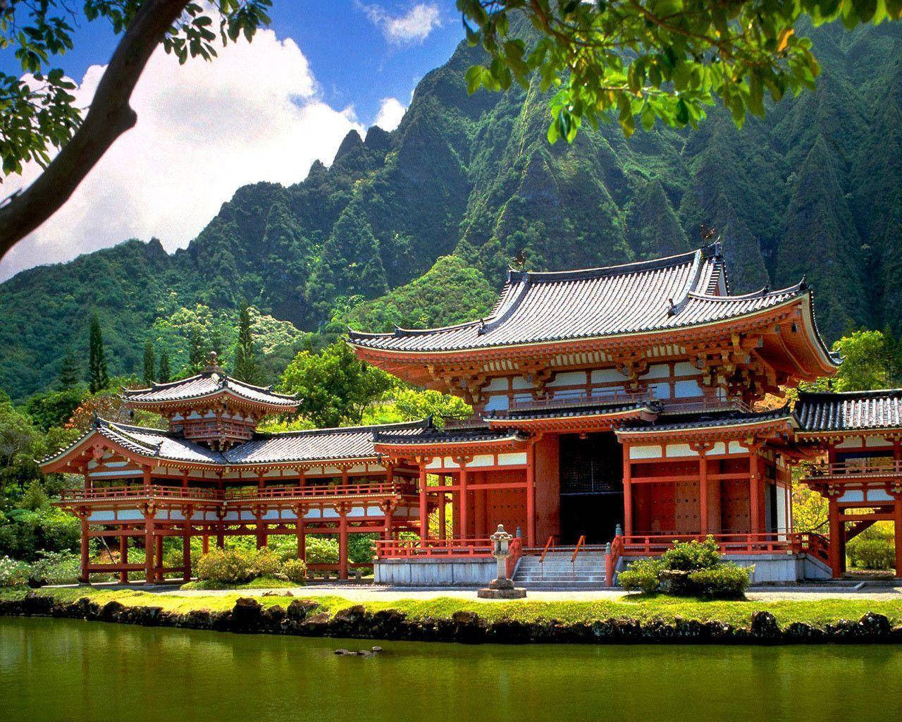 高清自然寺廟 壁紙 — 一座傳統紅色木造寺廟，擁有彎曲的屋頂，坐落在平靜的池塘旁，四周環繞著翠綠的山巒，在明亮的藍天下。