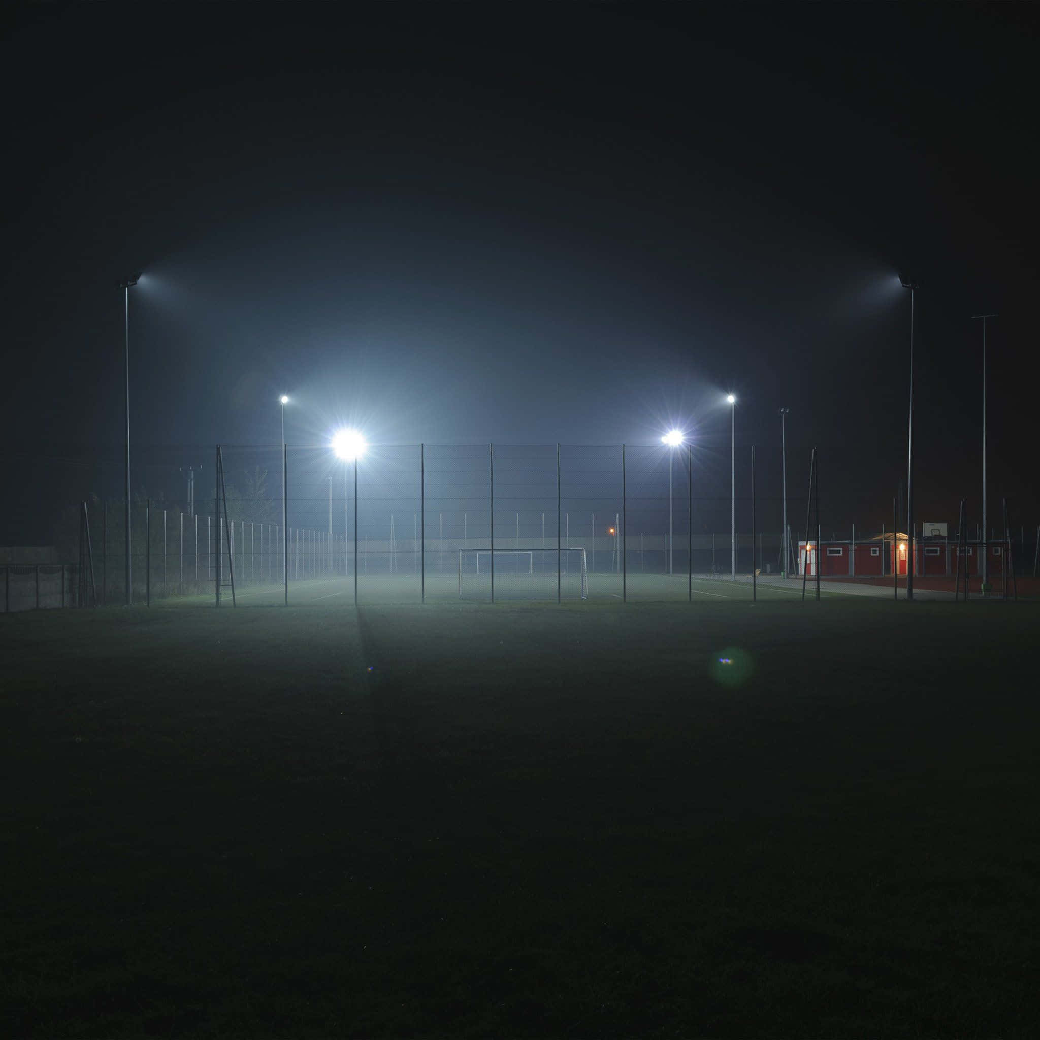 高燈柱足球場背景 壁紙 — 夜晚有霧的足球場被明亮的體育場燈光照亮，投射出長長的陰影和鏡頭光暈。
