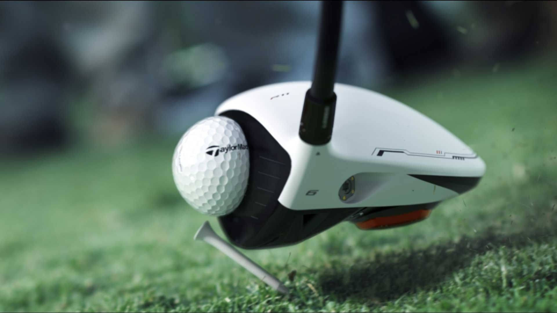 高爾夫揮桿慢動作畫面 壁紙 — TaylorMade 高爾夫球桿頭擊中綠色發球檯上的白色高爾夫球，草葉以慢動作飛散。