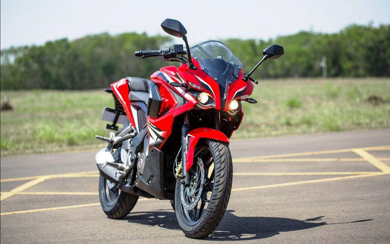 高畫質亮紅色 Pulsar RS200 機車 壁紙 — 一輛引人注目的紅色運動電單車，頭燈發亮，停泊在明亮的日光下的瀝青停車場。