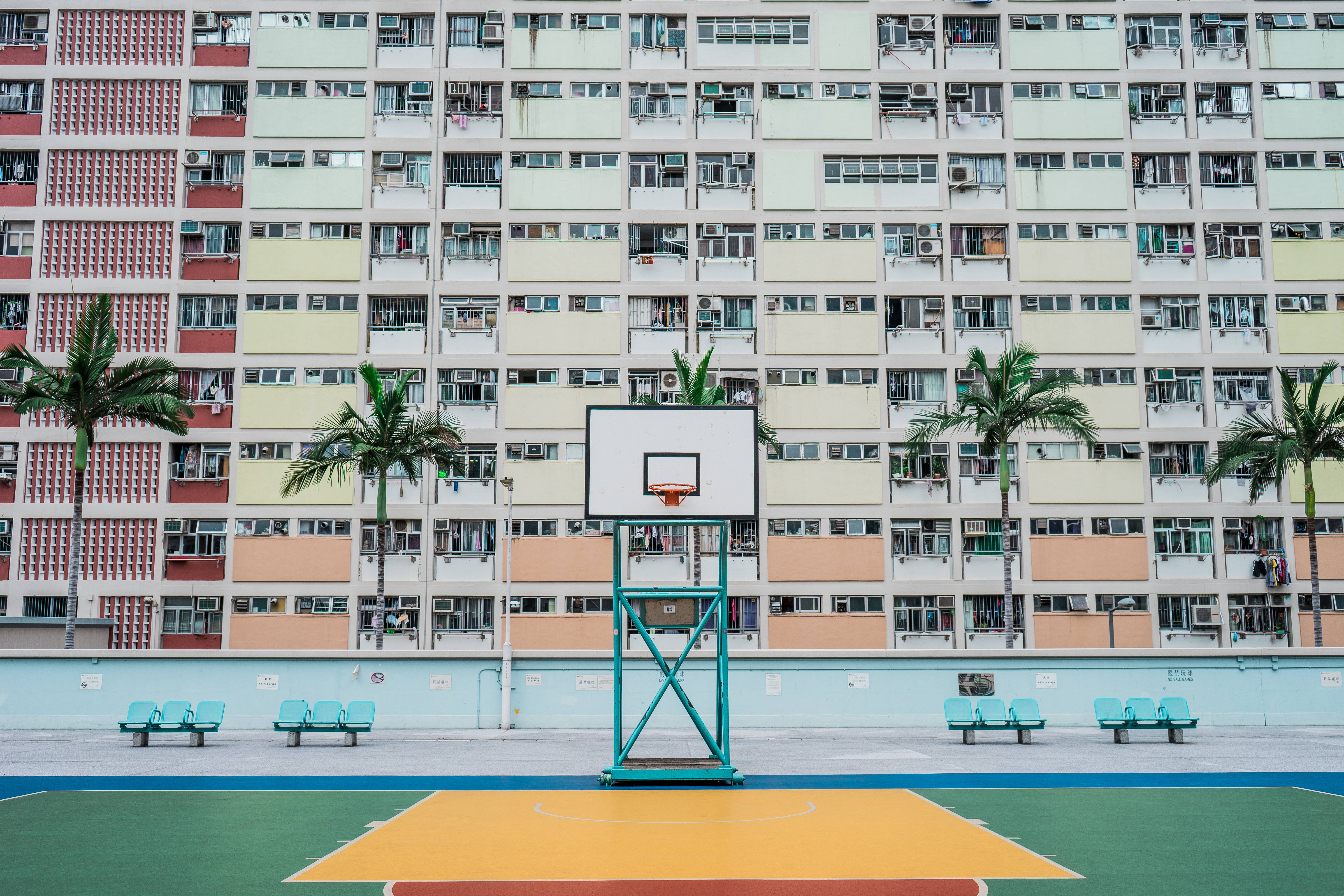 高畫質住宅區旁籃球場 壁紙 — 一個充滿活力的籃球場，帶有青綠色的籃框，矗立在一棟高聳、色彩繽紛的住宅大樓前，大樓兩旁種滿棕櫚樹。
