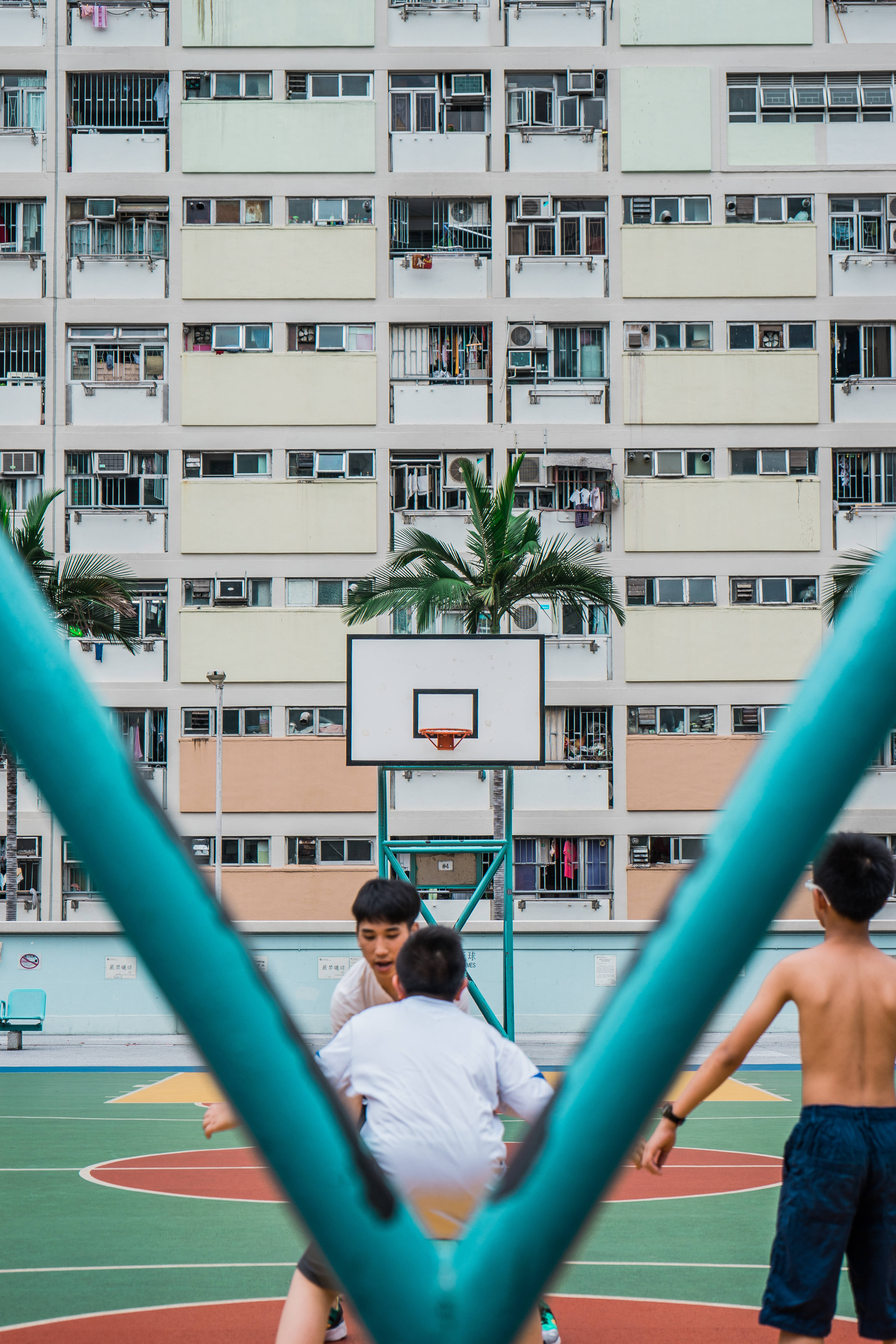 高畫質籃球與住宅區 壁紙 — 三個男孩在一個充滿活力的球場上打籃球，球場由青綠色的柱子框起，後面有一座密集的公寓大樓聳立。