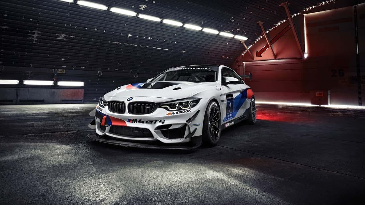 高畫質藍色寶馬 壁紙 — 一輛帶有藍色、紅色和黑色塗裝的白色BMW M4 GT4賽車停放在昏暗的工業車庫中，在明亮的燈光下。
