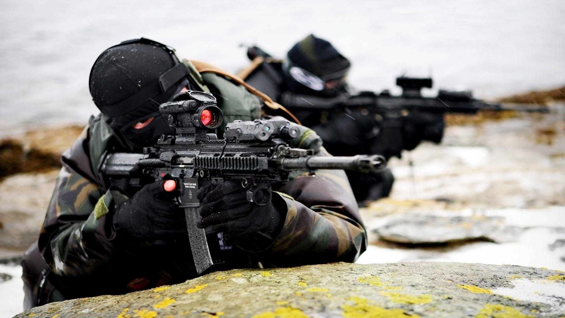 高畫質軍用瞄準鏡槍械 壁紙 — 兩名身穿迷彩裝備的士兵在陰天嘅雪岸上，喺岩石後面用步槍瞄準。