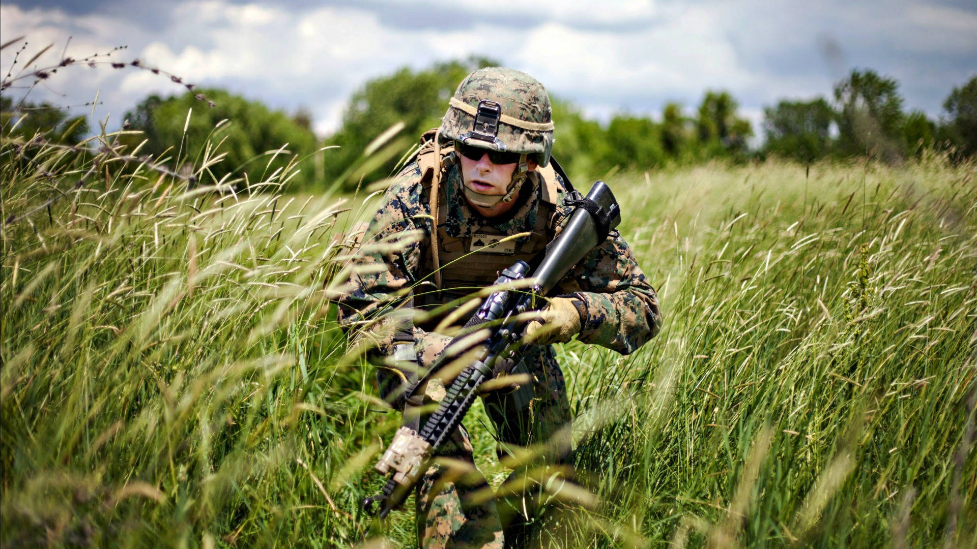 高畫質軍隊白日潛行 壁紙 — 一名身穿迷彩裝備的士兵蹲在高高的綠草中，手持步槍，天空多雲。
