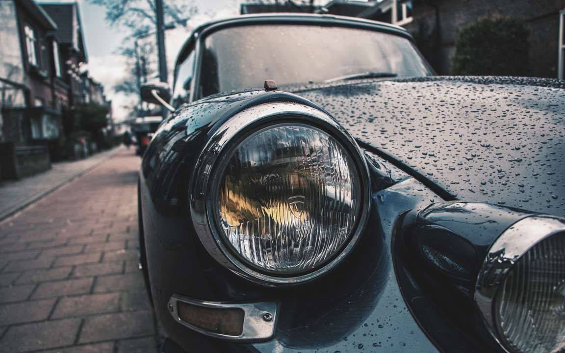 高畫質雨中車頭特寫 壁紙 — 一輛復古黑色汽車的車頭燈特寫，引擎蓋上閃爍著雨滴，停泊在濕漉漉的磚砌街道上。