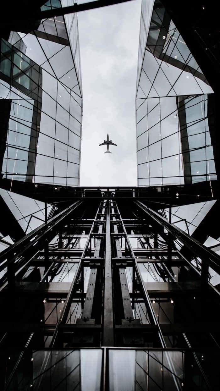 高空飛行 壁紙 — 一張低角度黑白照片，拍攝飛機在擁有反光外牆的 towering 玻璃摩天大樓之間飛行。