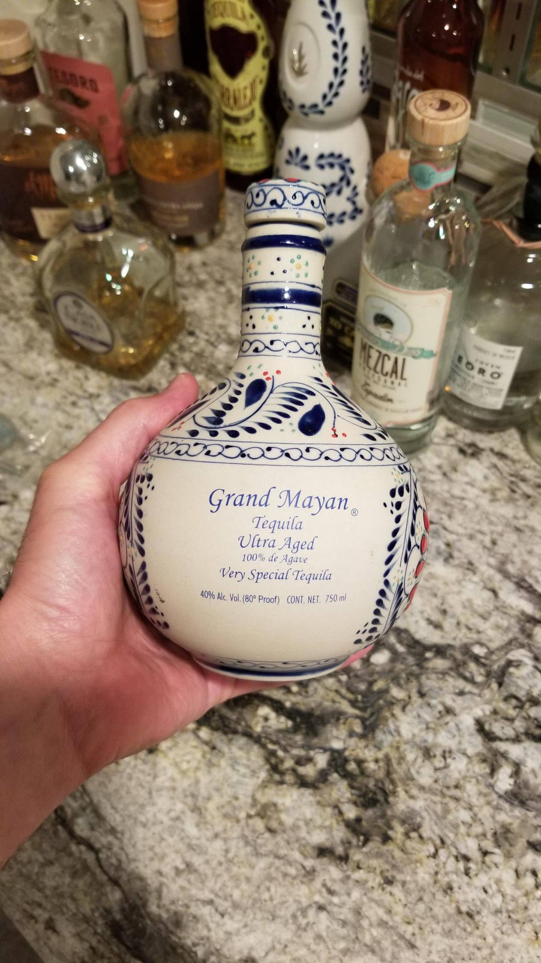 手拿著一個裝飾性龍舌蘭酒瓶，放在廚房櫃檯上，旁邊有其他酒瓶。