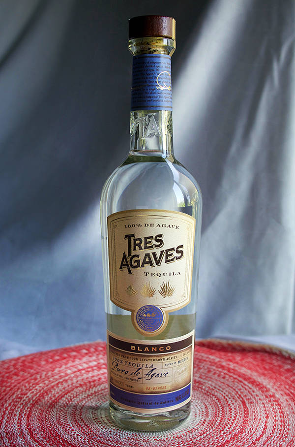 一個透明的龍舌蘭酒瓶，標有 Tres Agaves Blanco，放在紅色紋理墊上，背景是灰色布料。