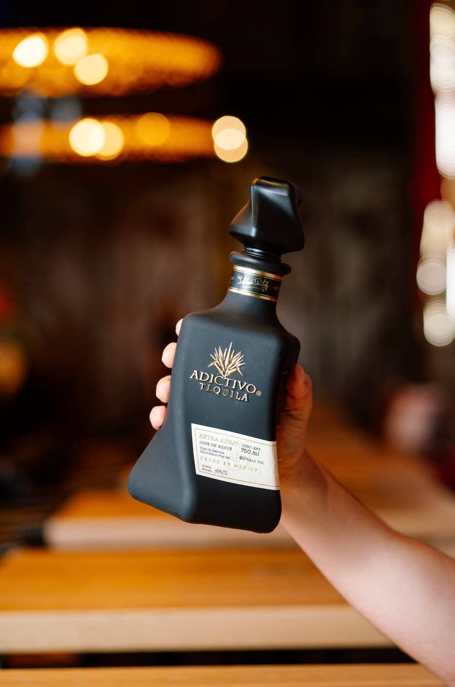 高級Adictivo龍舌蘭酒瓶 壁紙 — 一隻手拿著一瓶時尚的黑色Adictivo Tequila，背景是溫暖燈光的酒吧，帶有模糊的金色散景燈光。