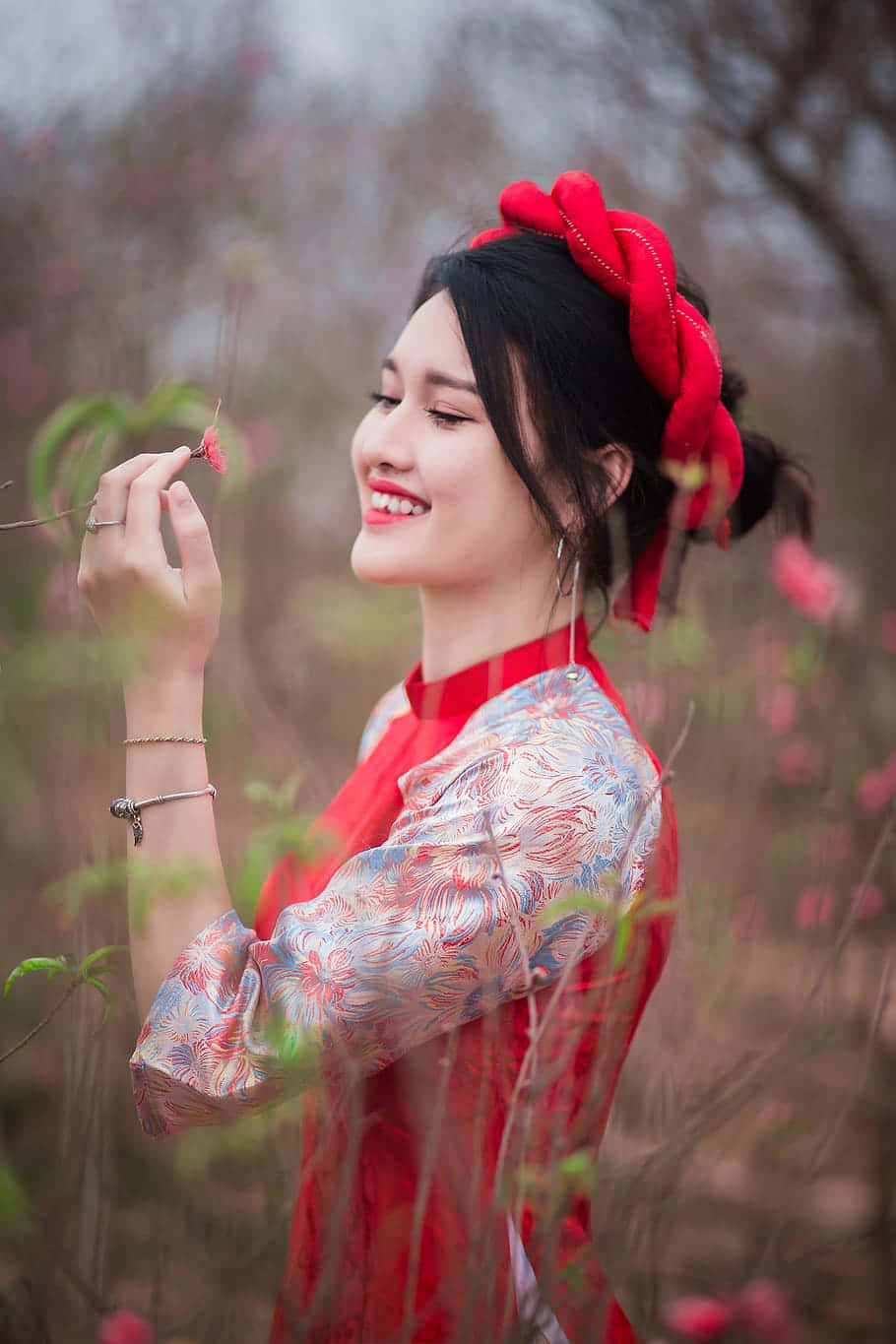 高級時尚連衣裙於優雅場景 壁紙 — 一位微笑的女子身穿紅色圖案連衣裙和相配的頭飾，在柔和的春日花園中輕輕捧著一朵櫻花。