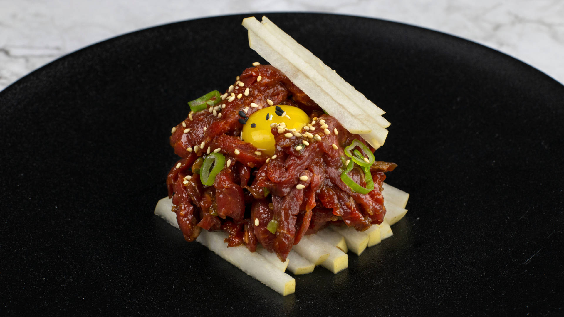 高級牛排塔塔配香料與配菜 壁紙 — 一個精心擺盤的菜餚，以黑盤盛載辛辣醃製牛肉，上面放有生蛋黃和芝麻。