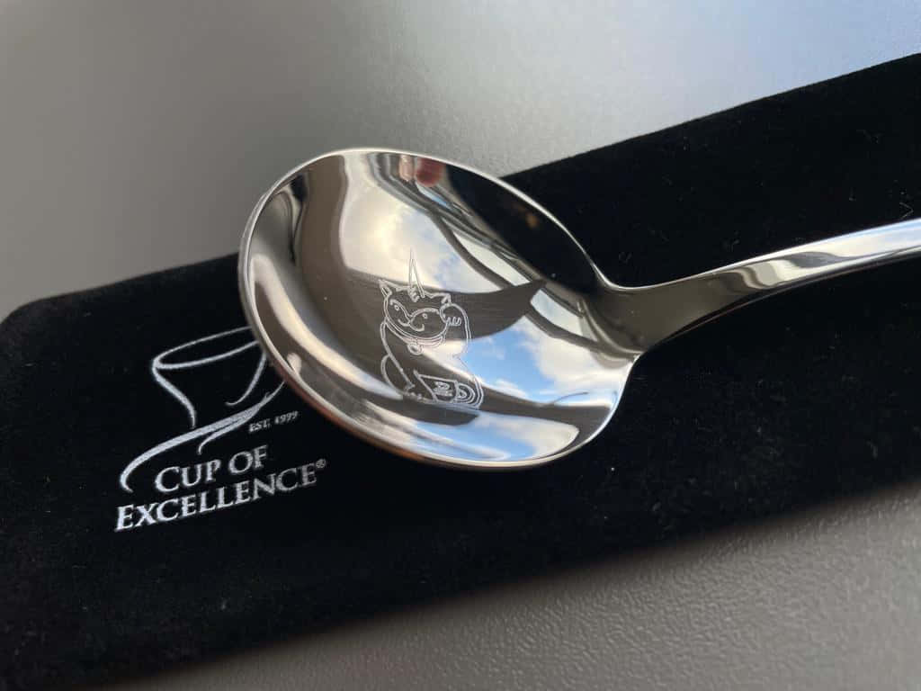 高級銀湯匙 壁紙 — 一隻閃亮的銀匙，上面刻有貓圖案，放在印有「Cup of Excellence」品牌的黑色天鵝絨袖套上。