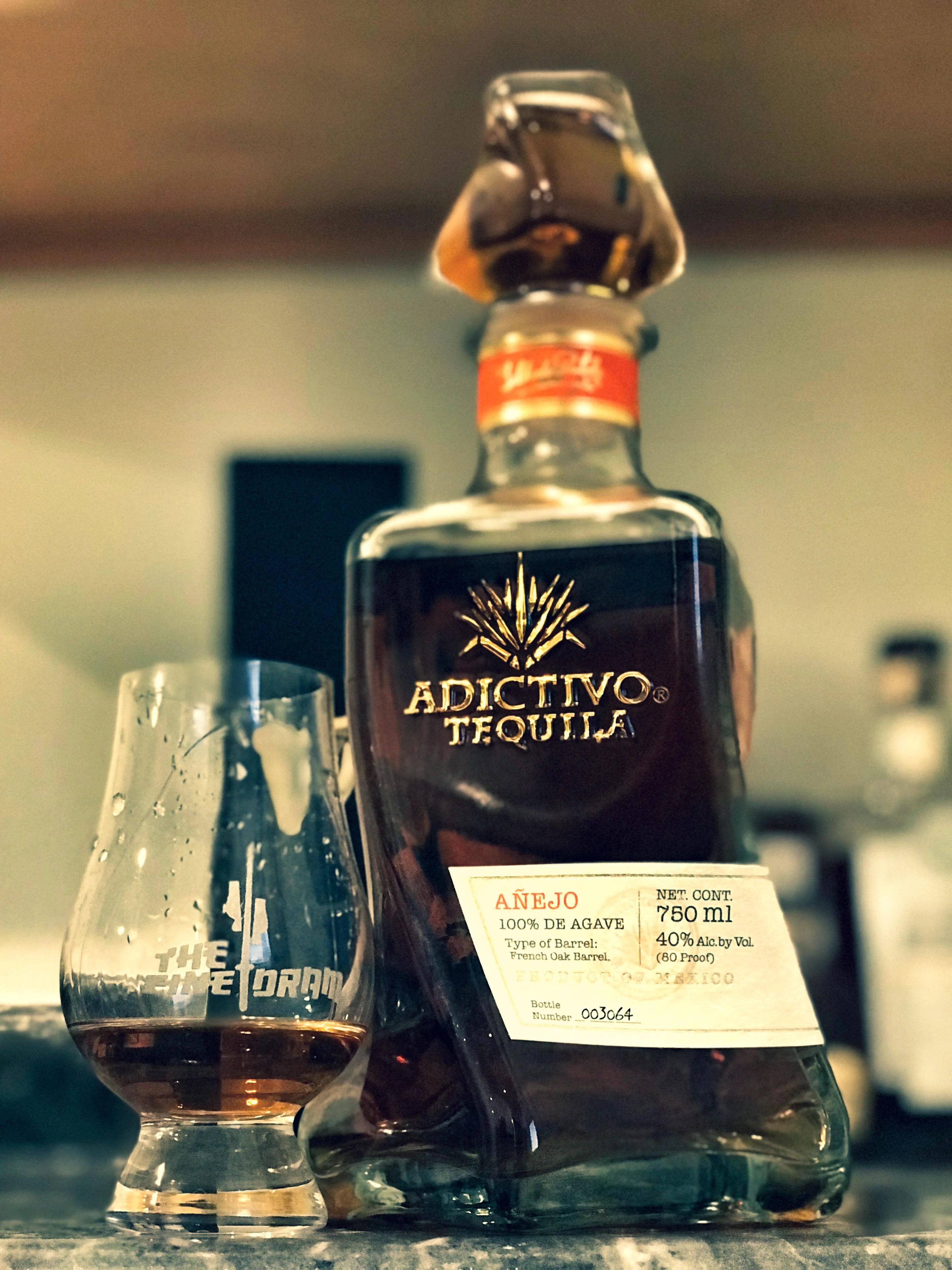 高級阿迪克蒂沃黑龍舌蘭特級陳年瓶 壁紙 — 一瓶 Adictivo Añejo 龍舌蘭酒放在吧檯上，旁邊有一個品酒杯，環境燈光溫暖。