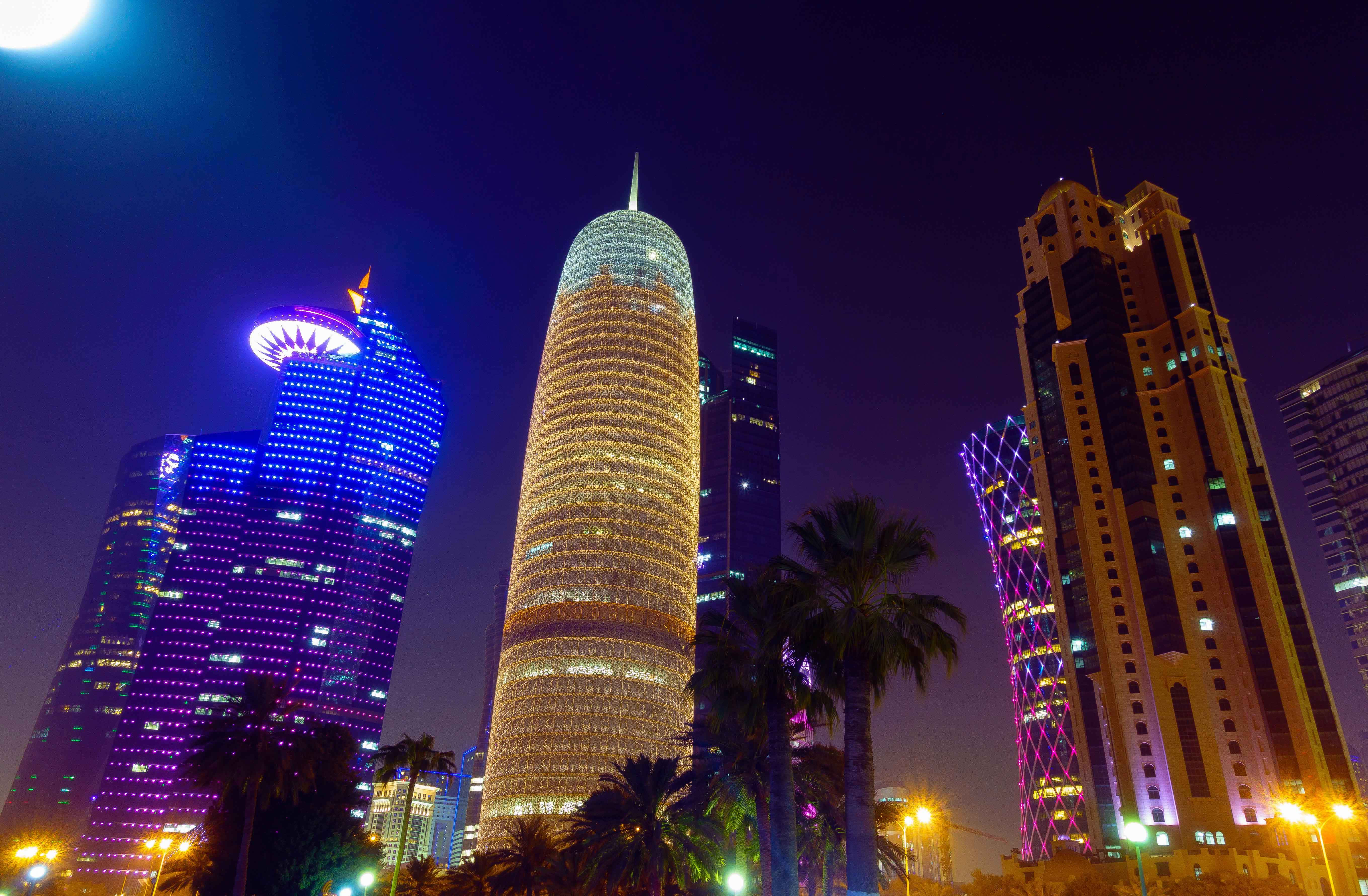 Qatar 桌布 壁紙 — 高聳的摩天大樓在深邃的夜空中閃爍著鮮豔的霓虹燈光，棕櫚樹在下方形成剪影。