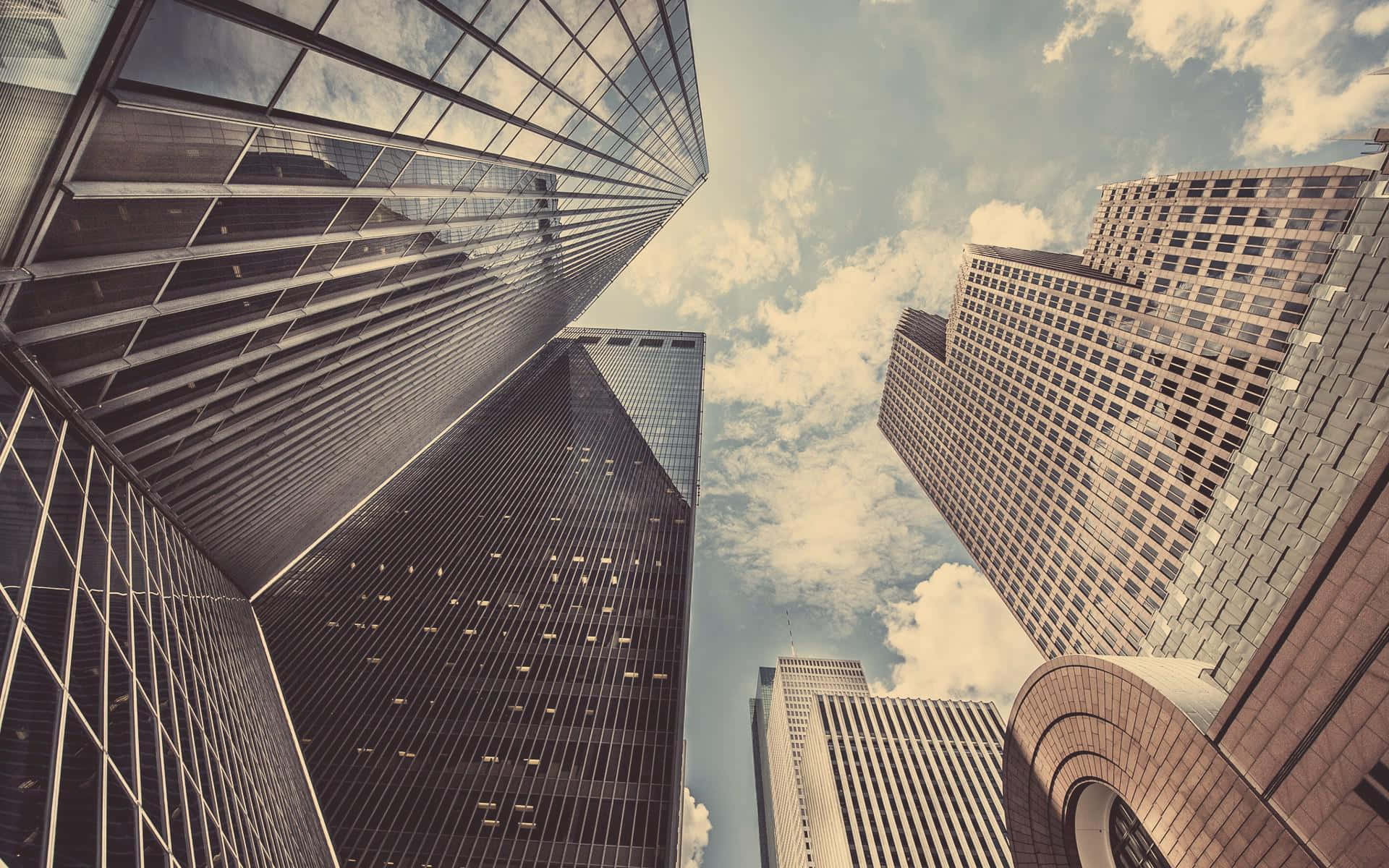 Buildings 桌布 壁紙 — 高聳的玻璃和鋼鐵摩天大樓在局部多雲的天空下向上匯聚，形成一個戲劇性的低角度城市景觀。