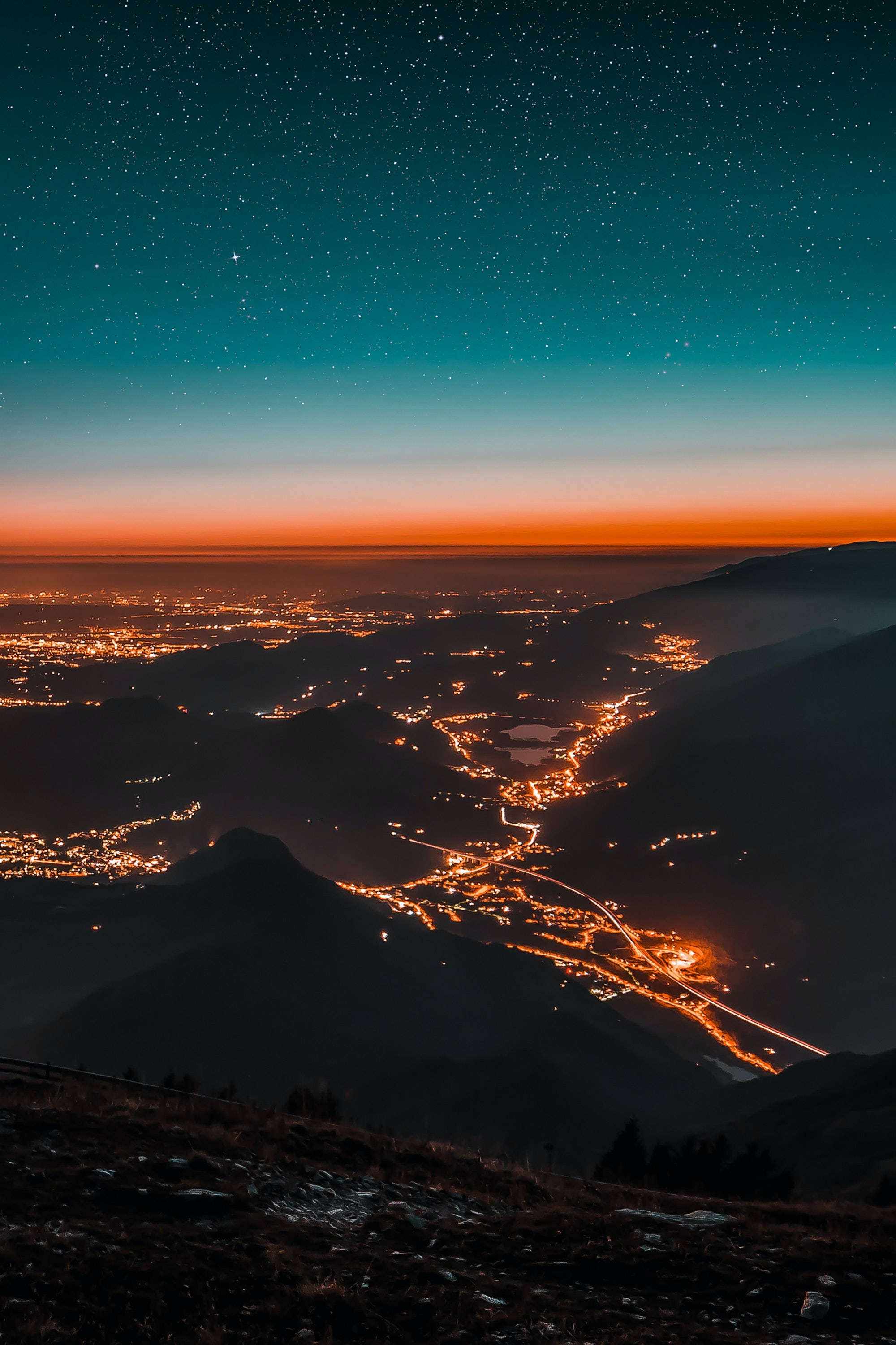 高角度城市夜景 壁紙 — 星空夜空在山谷小鎮上空發光，橙色燈光蜿蜒穿過黑暗的山脈。