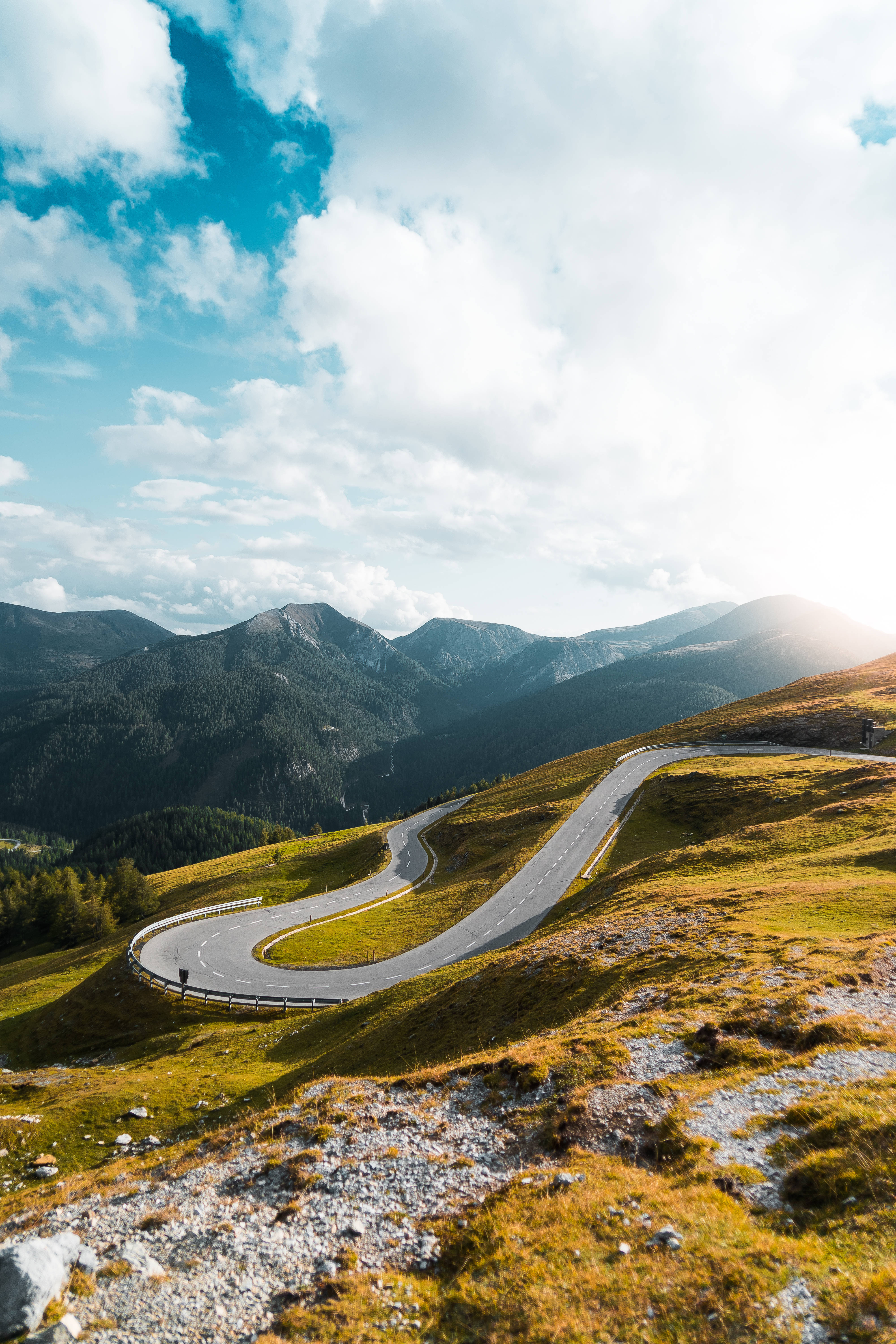 高解析度 iPhone 山路風景 壁紙 — 一條蜿蜒的山路在陽光明媚的草地丘陵間蜿蜒曲折，上方是明亮的藍天和蓬鬆的白雲。
