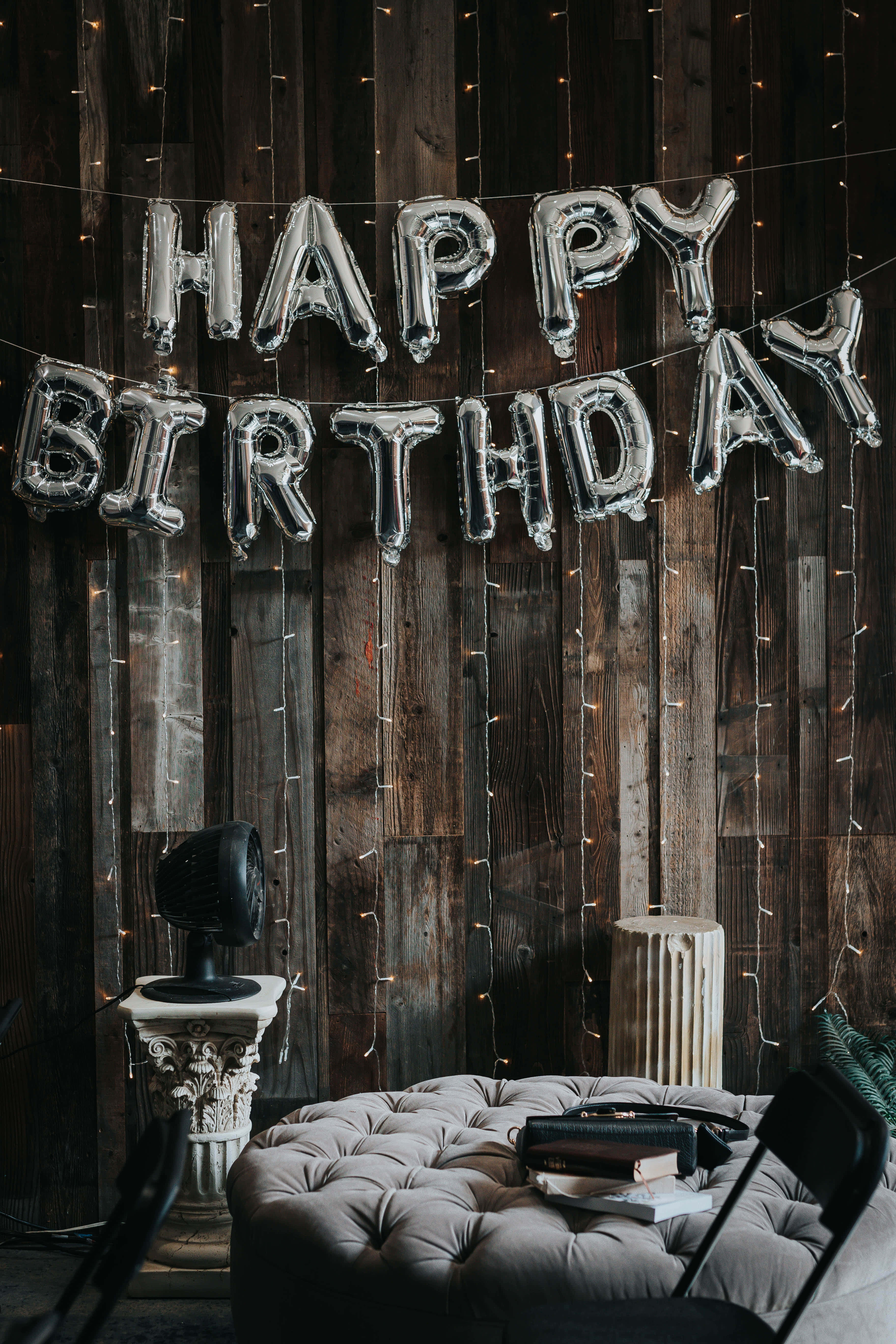 高解析度快樂生日，慶祝特別的日子 壁紙 — 銀色 'HAPPY BIRTHDAY' 氣球掛在質樸的木牆上，牆上裝飾著燈串，下方是簇絨灰色軟凳。