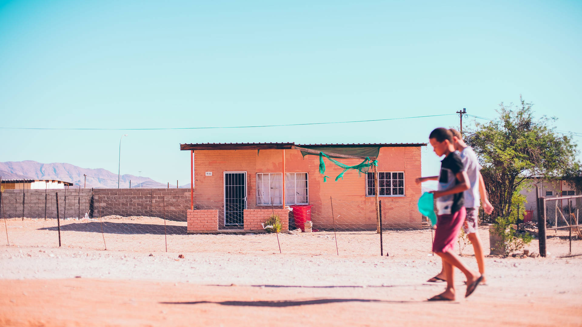 兩個孩子赤腳走過一間桃色房子，在乾燥的庭院裡，陽光明媚。 — 高解析度桌面 壁紙